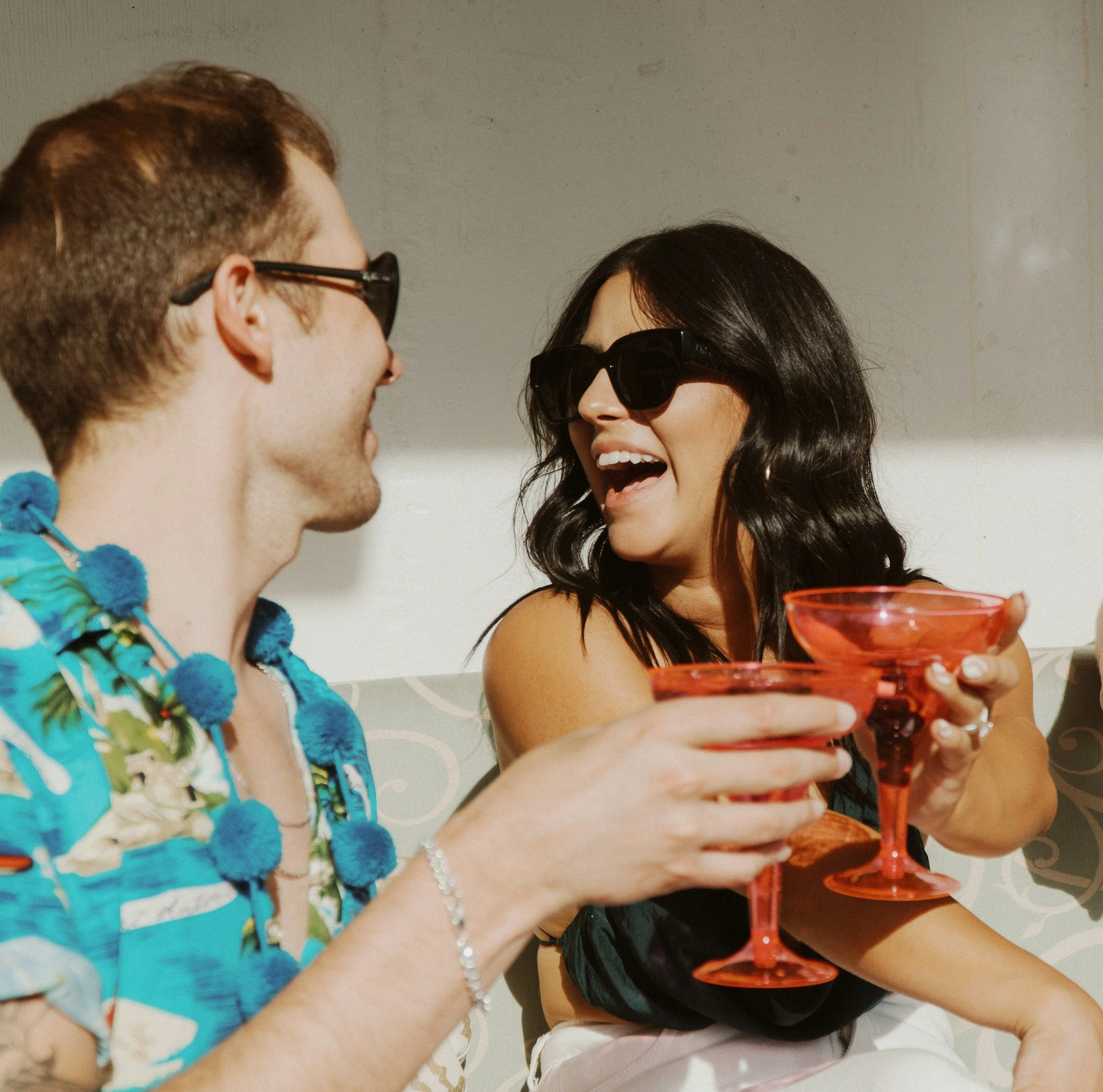 Two women wearing sunglasses face each other, smiling, with a woman on the right holding a layered drink in a red glass. The woman on the left has short brown hair and is wearing a tropical shirt with blue pom-pom accessories. The woman on the right has black hair styled in loose waves and is wearing a white top.