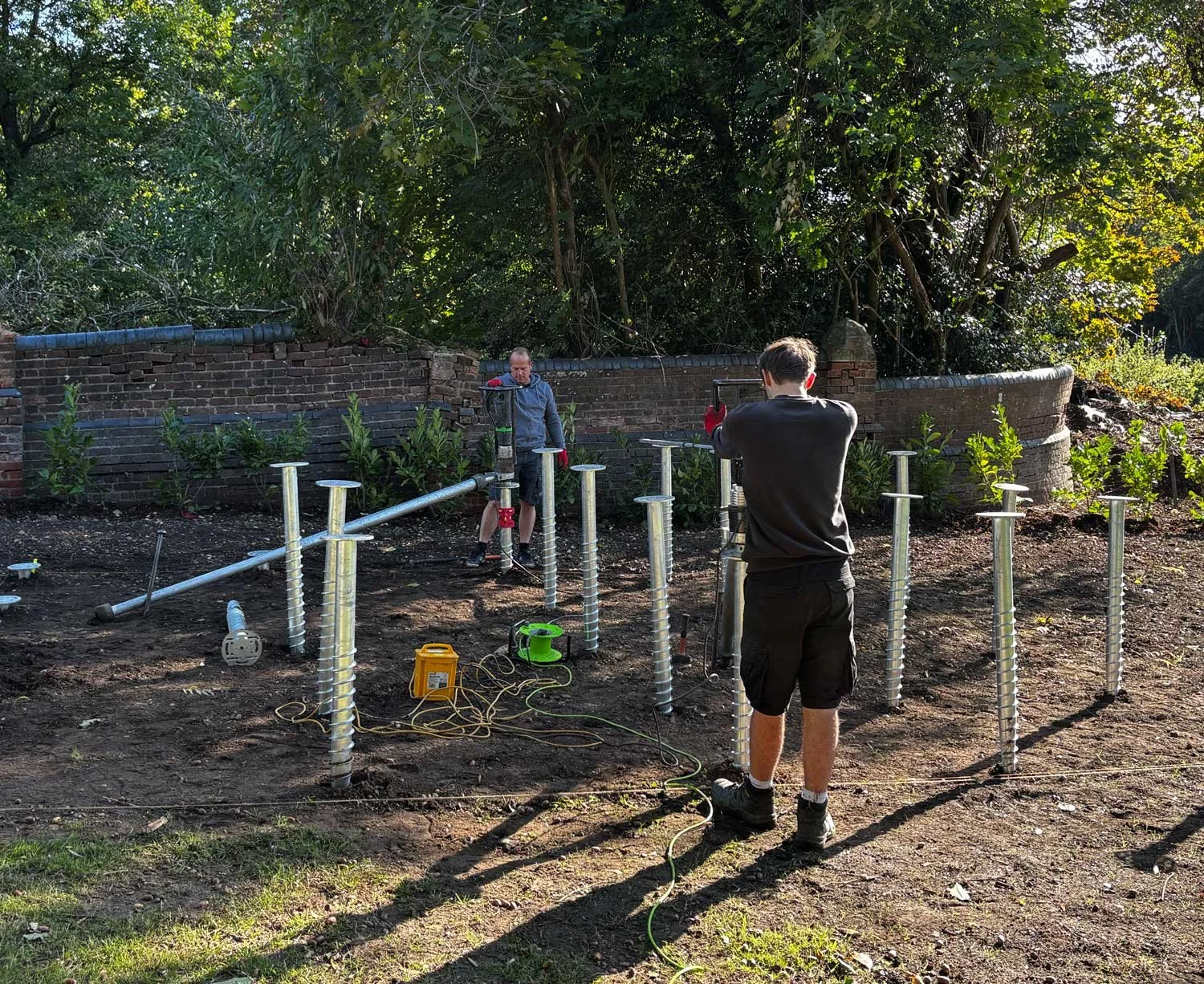Two young men working on a construction site outdoors, installing large metal screw piles into the ground. One man faces away from the camera, the other is positioned near a brick wall with trees behind them, illuminated by sunlight.