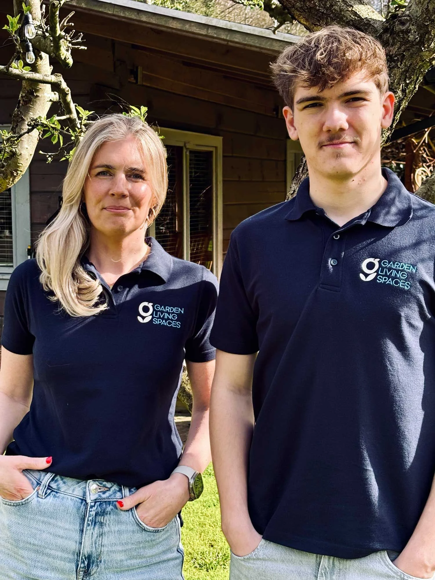 Two people standing outdoors near a tree, wearing black shirts with the logo 'Garden Living Spaces'.