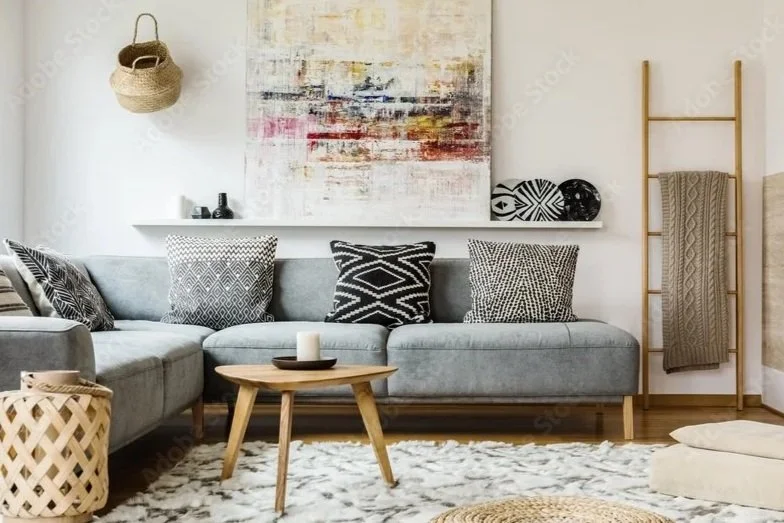 Living room with a gray sectional sofa decorated with patterned black and white pillows, a small wooden coffee table with a candle, a white textured rug, a wall shelf with black and white decorative items, an abstract painting, a woven basket hanging on the wall, a wooden ladder with a throw blanket, and a floor cushion.