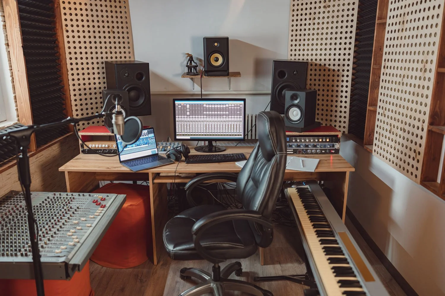 Music studio with equipment including large monitors, audio mixer, microphone, keyboard, and computer, with soundproofing walls.