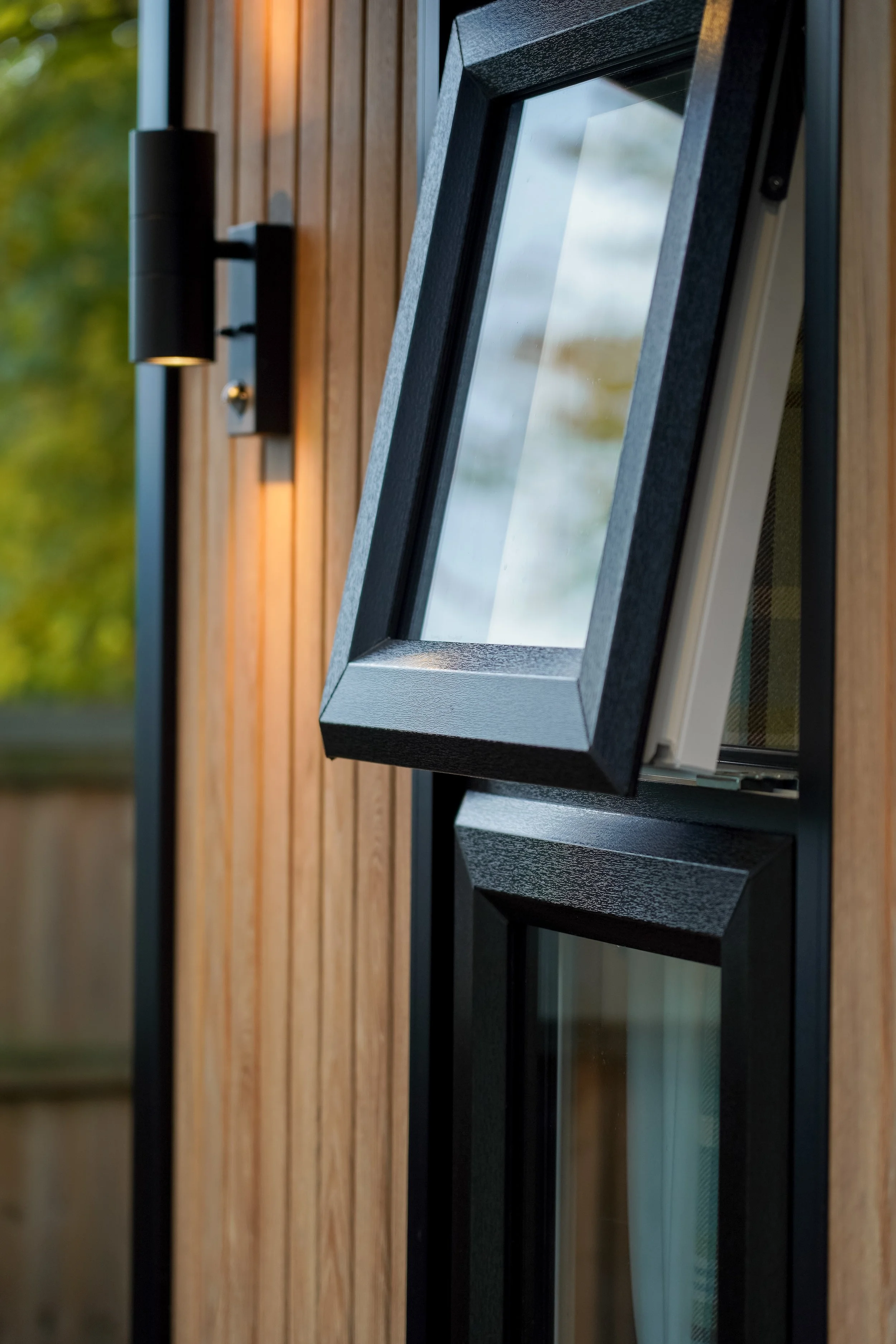 A close-up of a window with a black frame, set in a wooden wall with vertical planks, and a black exterior wall lamp beside it. The window reflects the sky and clouds.