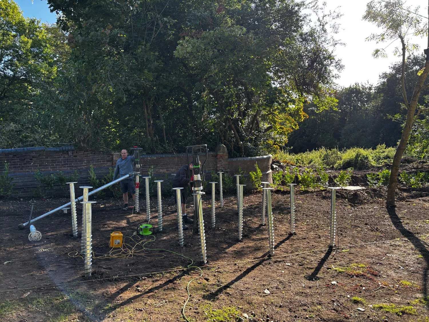 People installing large metal screws into the ground in a garden, with a brick wall and trees in the background.