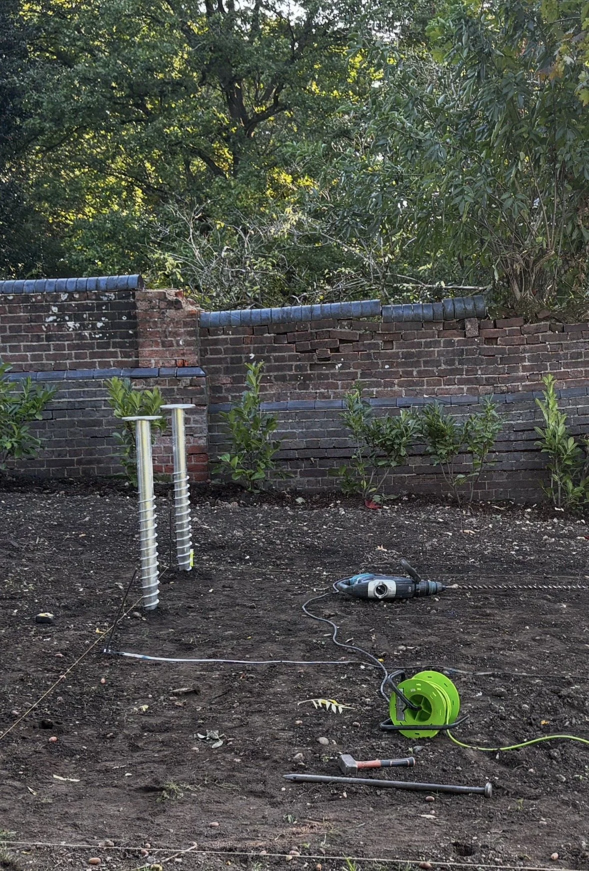 A garden with a partially built brick wall, plants along the base, tools on the ground including a drill and a green extension cord reel, and a background of tall trees and shrubs.