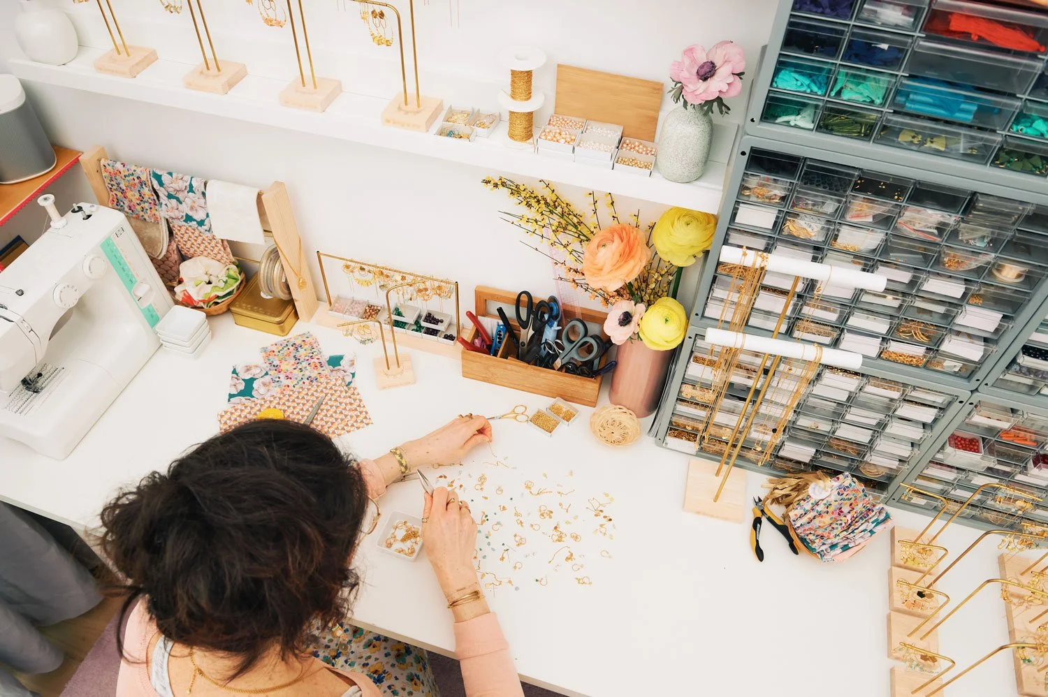 A woman working at a craft or jewelry station with sewing supplies, scissors, and fabric on a white table, surrounded by boxes of beads and jewelry findings, and decorative flowers in vases.
