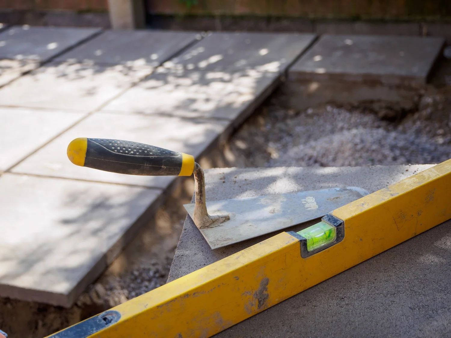 Close-up of a yellow spirit level and a trowel resting on a concrete surface during a tile installation at a construction site.