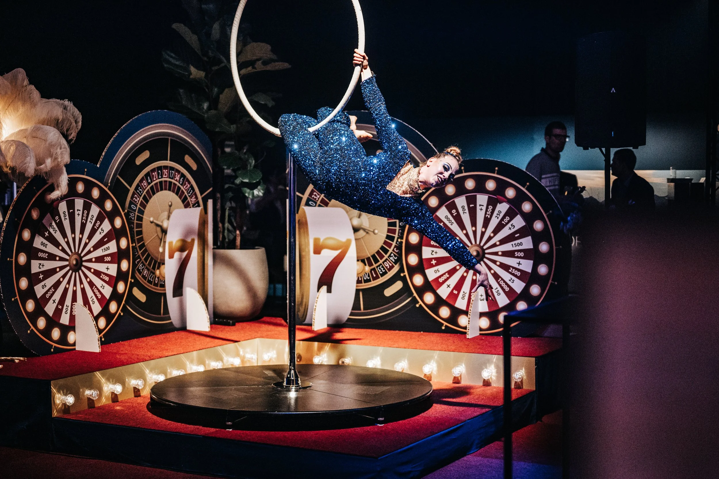 A female acrobat in a sparkly blue jumpsuit performing a flying trapeze act in a show. She is hanging upside down by her legs from a white hoop, with her arms extended. The background features roulette wheels and bright lights, creating a casino-like atmosphere.