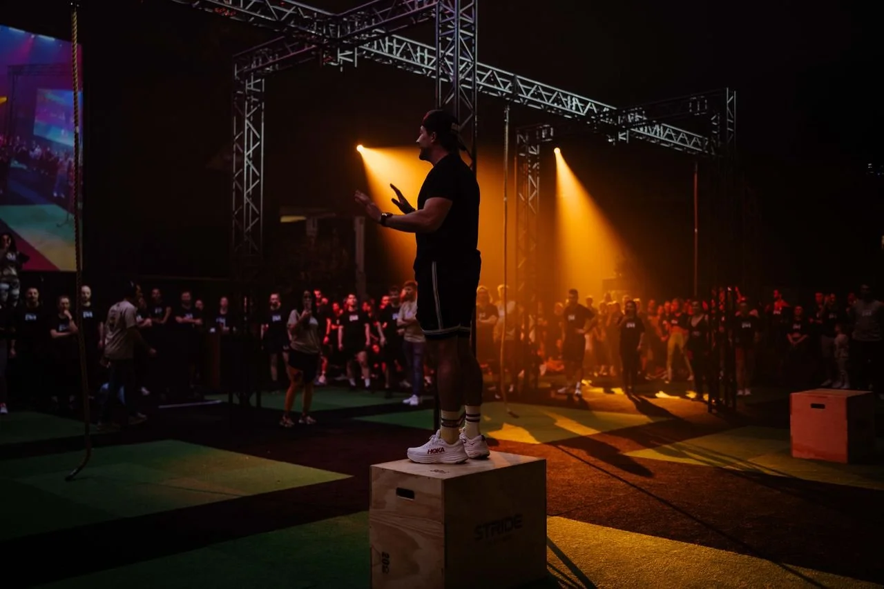 A man standing on a wooden box, speaking or explaining in front of a large crowd inside a dark gym or fitness center with orange lighting and a high metal frame structure overhead. The crowd is gathered around, some watching and some engaged in activities.
