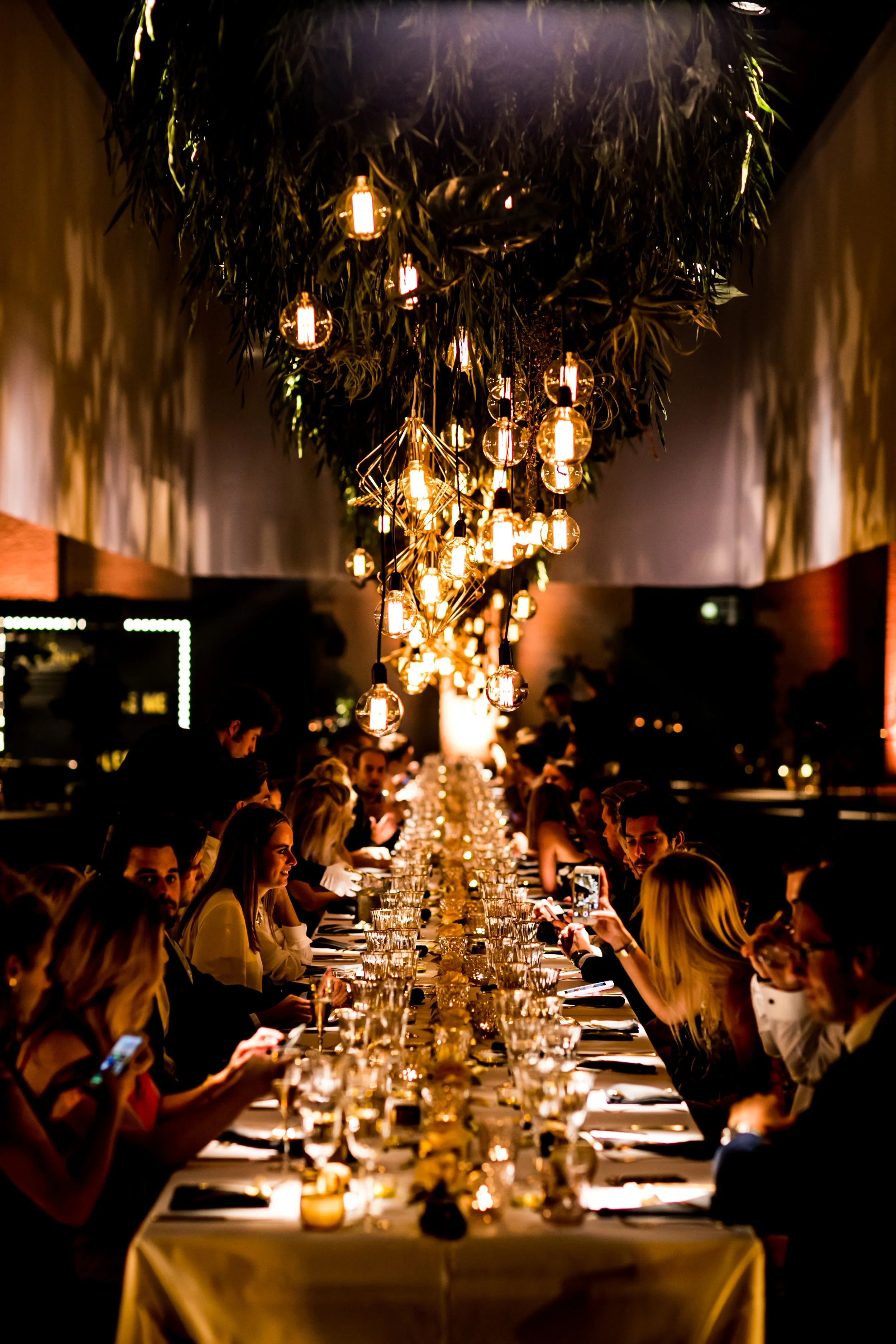 People dining at a long banquet table in a dimly lit restaurant with decorative hanging lights and lush greenery above.