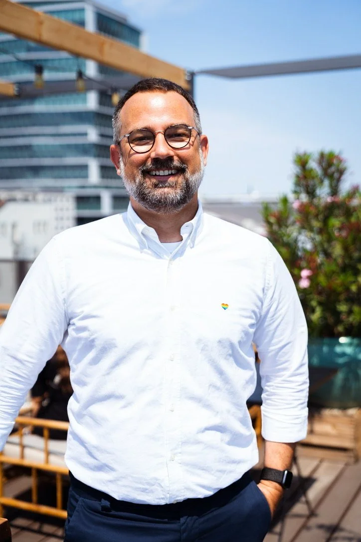A smiling man with glasses, a beard, and short dark hair wearing a white button-up shirt with a small rainbow heart logo on the chest, standing outdoors on a rooftop or terrace with city buildings and a blue sky in the background.
