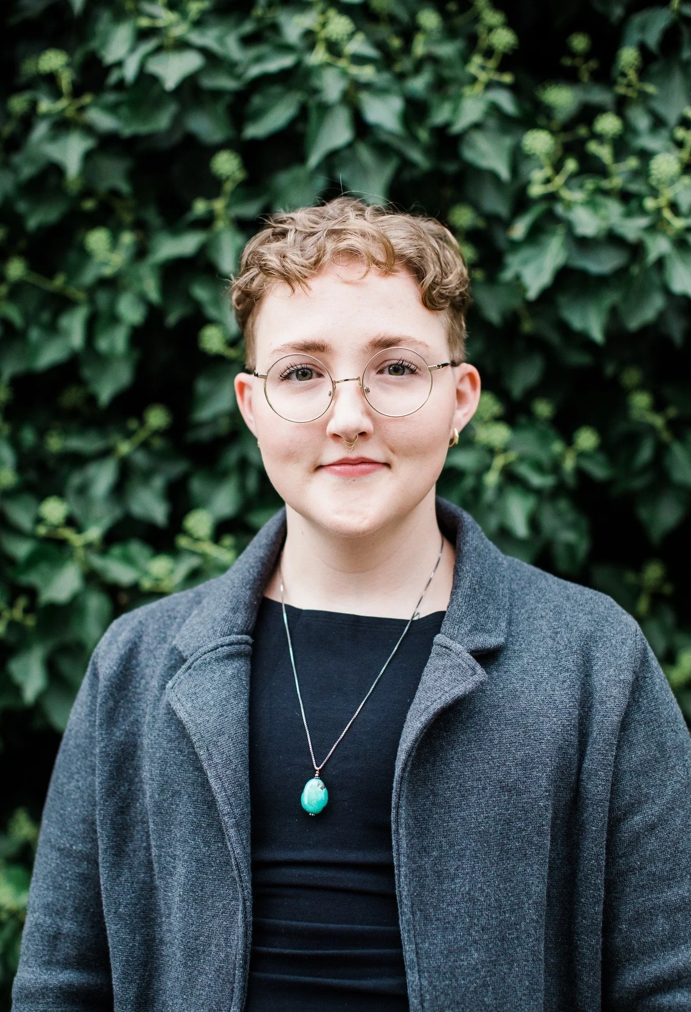 Portrait of a young person with short, curly hair wearing glasses, a septum piercing, a black shirt, and a gray coat, standing in front of green leafy bushes.