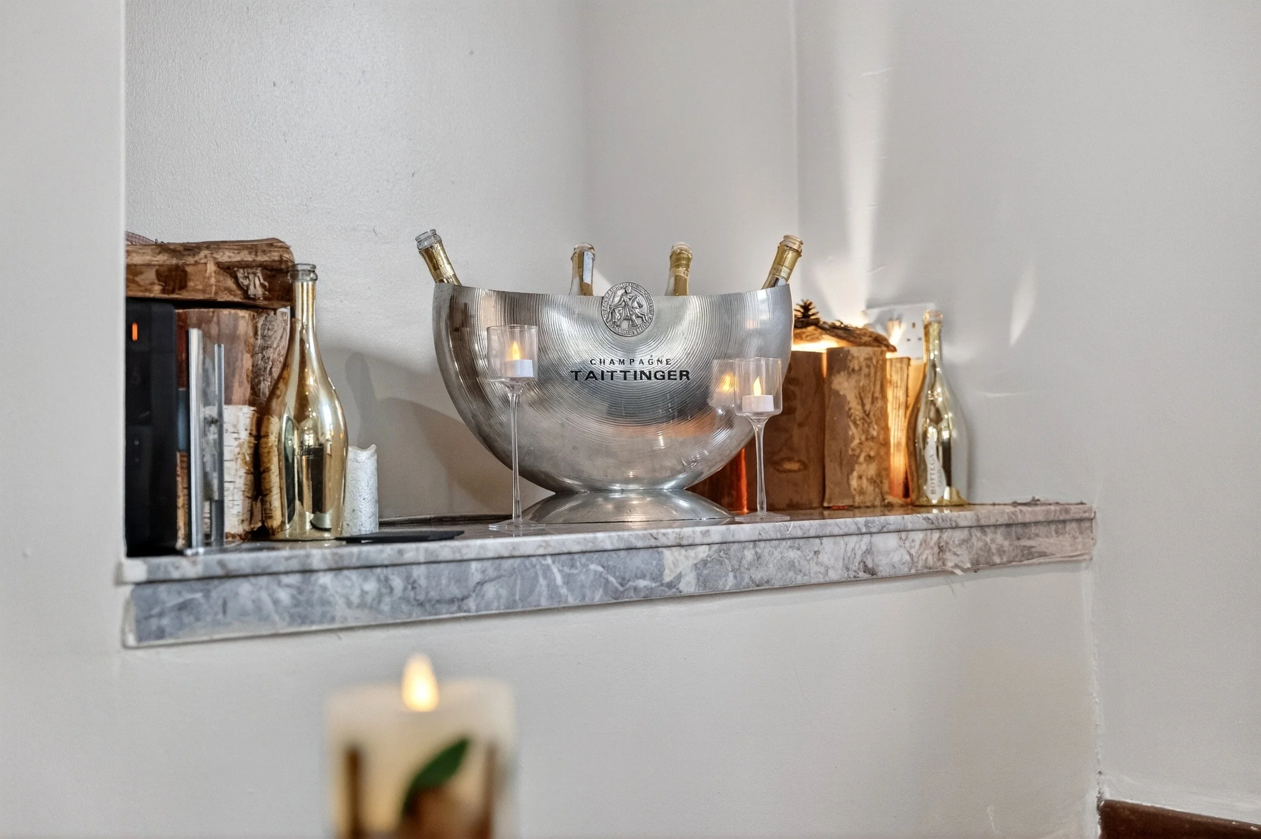 A decorative shelf displaying champagne bottles, a metal ice bucket, and lit candles, with a candle in the foreground.