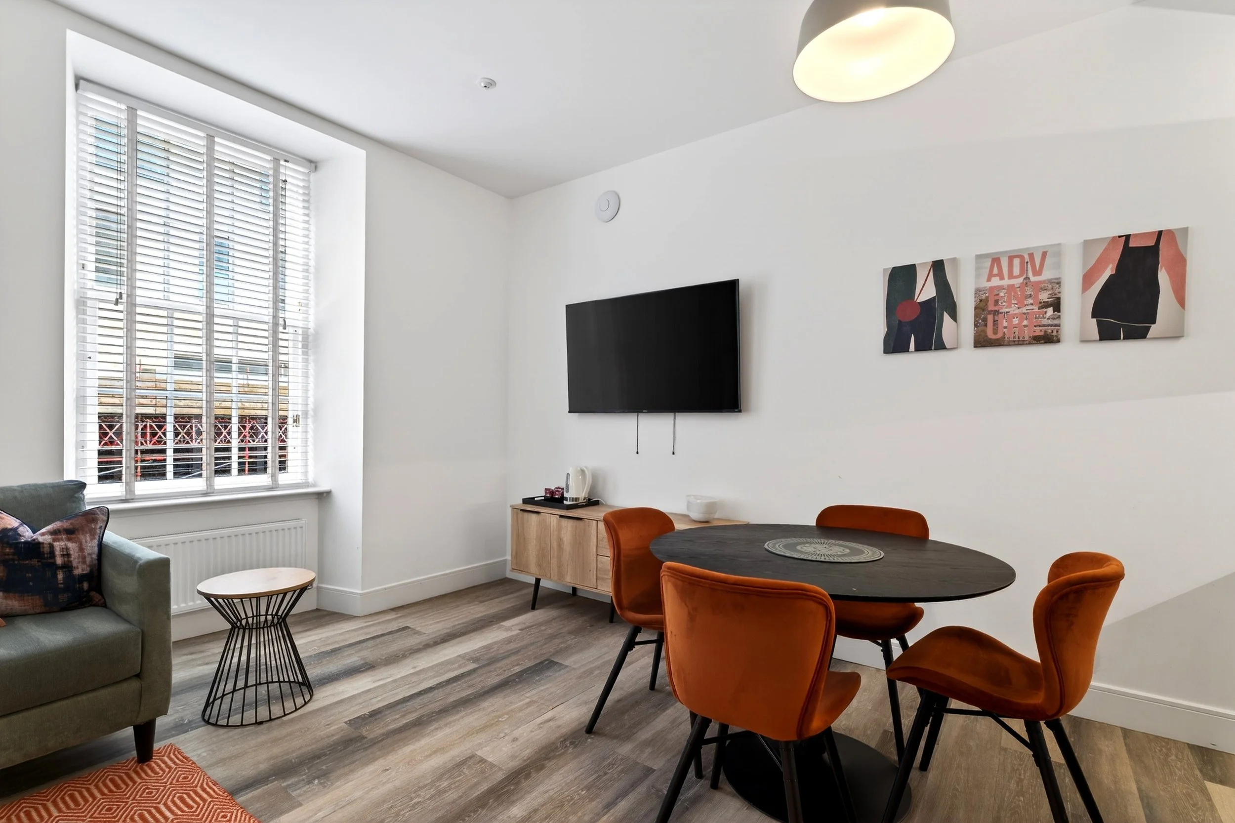 Modern living room with a window with white blinds, a wall-mounted TV, a wooden sideboard with a kettle and a bowl, a round black dining table with four orange chairs, and a colorful abstract wall art piece.