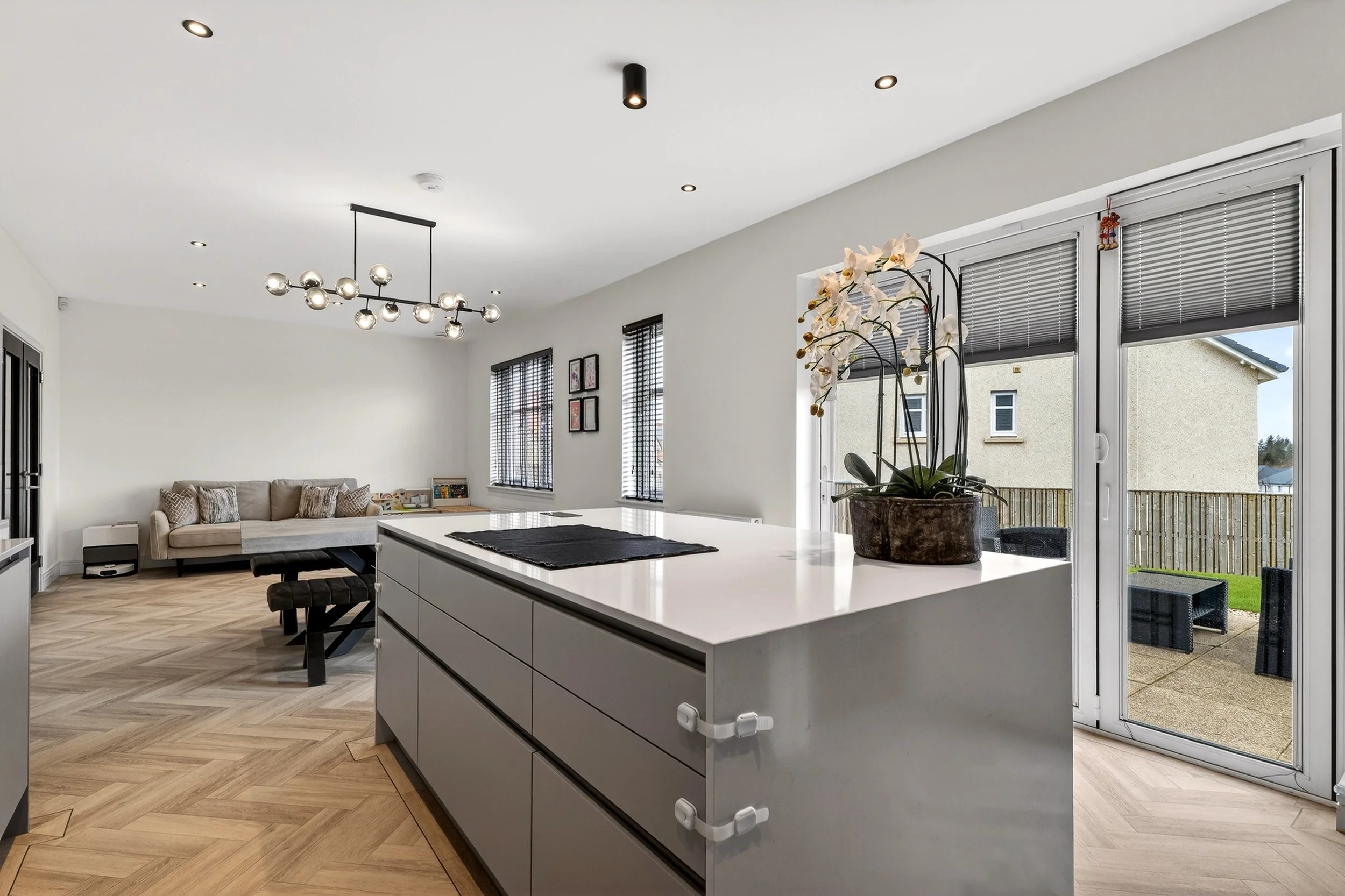 Modern open-concept living room and kitchen with white walls, hardwood flooring, large windows with blinds, a glass door leading to an outdoor patio, a beige sofa, dining table, decorative wall art, ceiling lights, and a potted orchid on the kitchen 