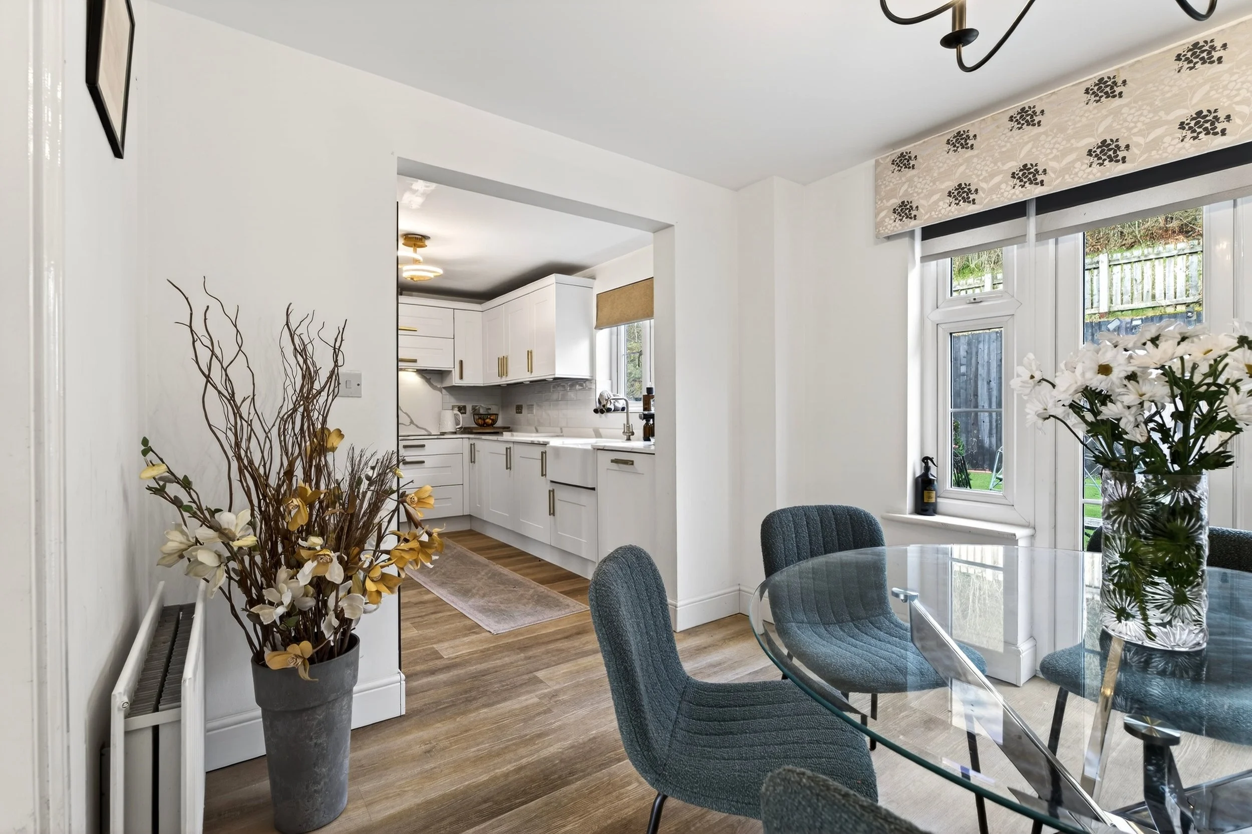 Modern dining room with a glass table, blue chairs, and a large floral centerpiece. Adjacent kitchen with white cabinets and gold handles, a window with a beige blind, and a doorway leading to the yard. Decor includes a tall vase with artificial flow