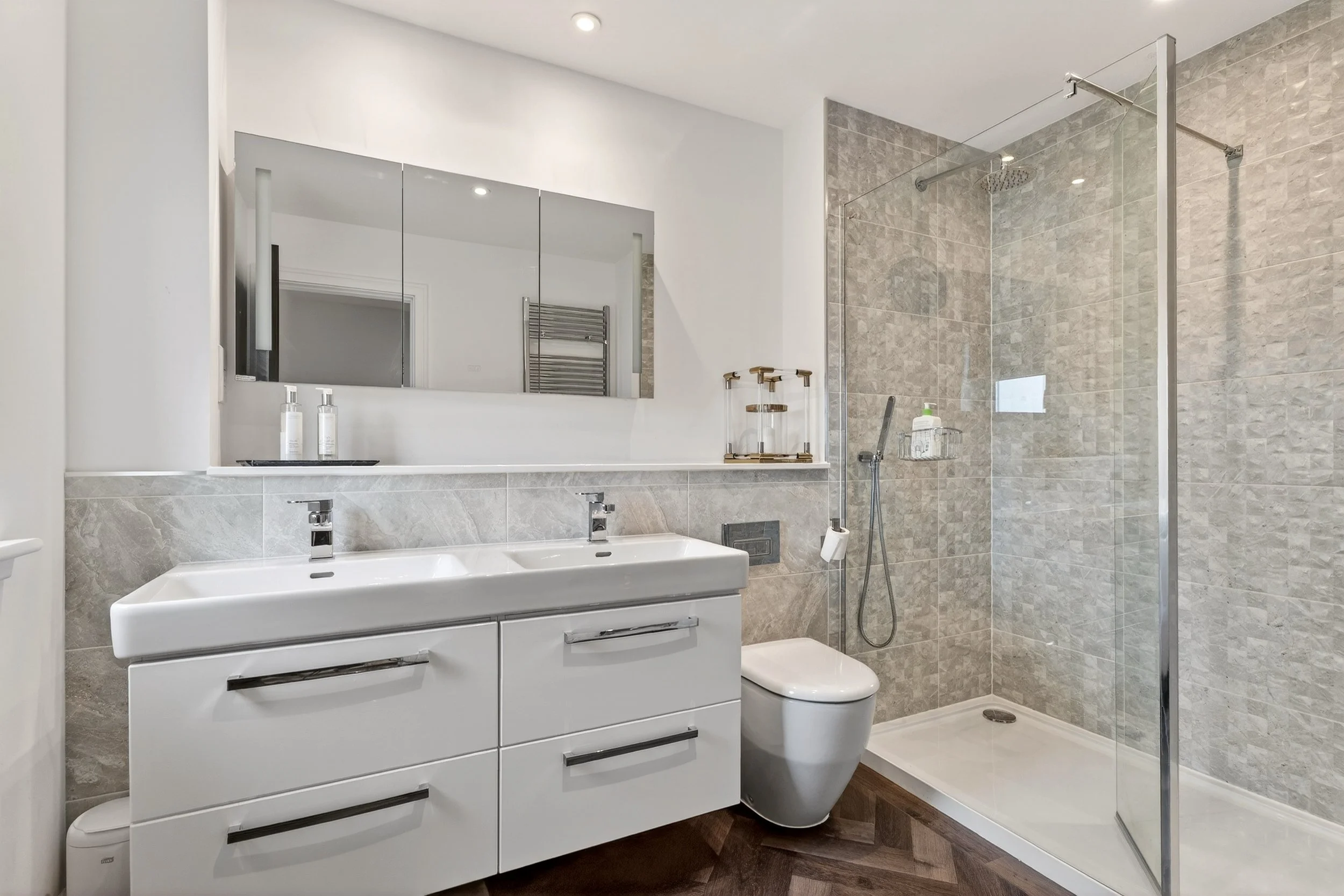 Modern bathroom with dual sinks, large mirror, wall-mounted toilet, and a glass-enclosed shower with beige tile walls.