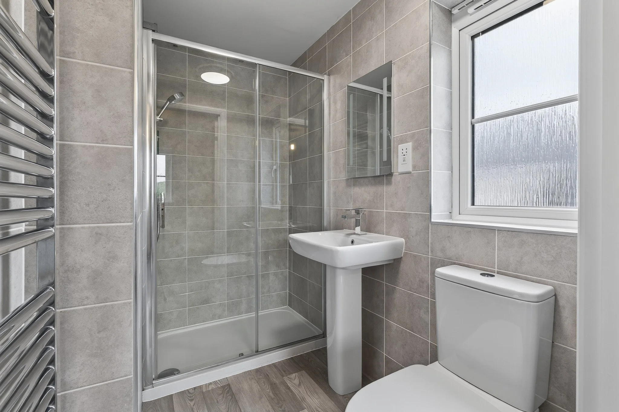 Bathroom with a glass shower enclosure, white sink on a pedestal, toilet, window with frosted glass, and beige tiled walls.