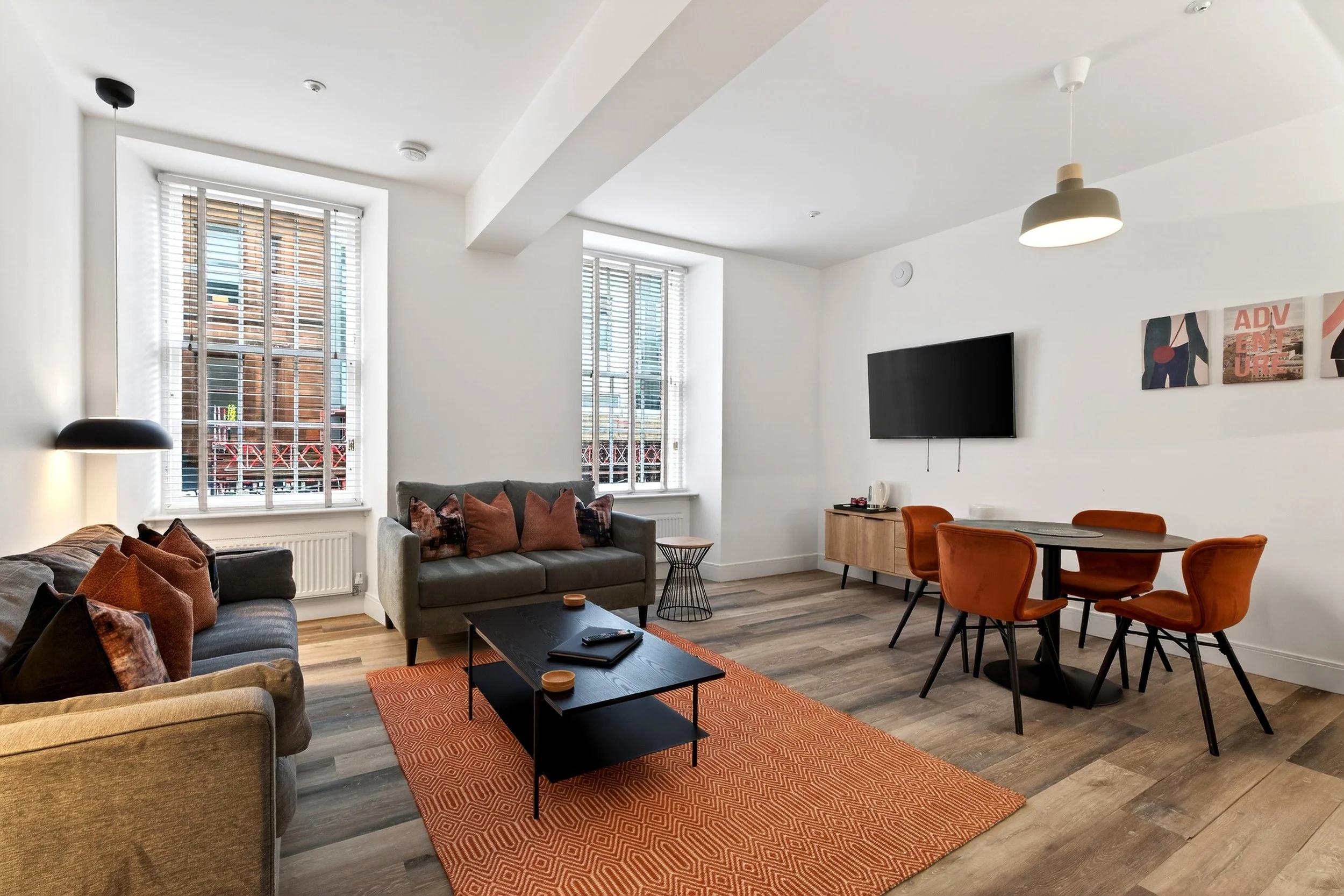 Living room with two gray sofas, one dark and one light, and a black coffee table on an orange rug. There are several cushions on the sofas, a small side table, and a wall-mounted television. Two large windows with white blinds allow natural light, a