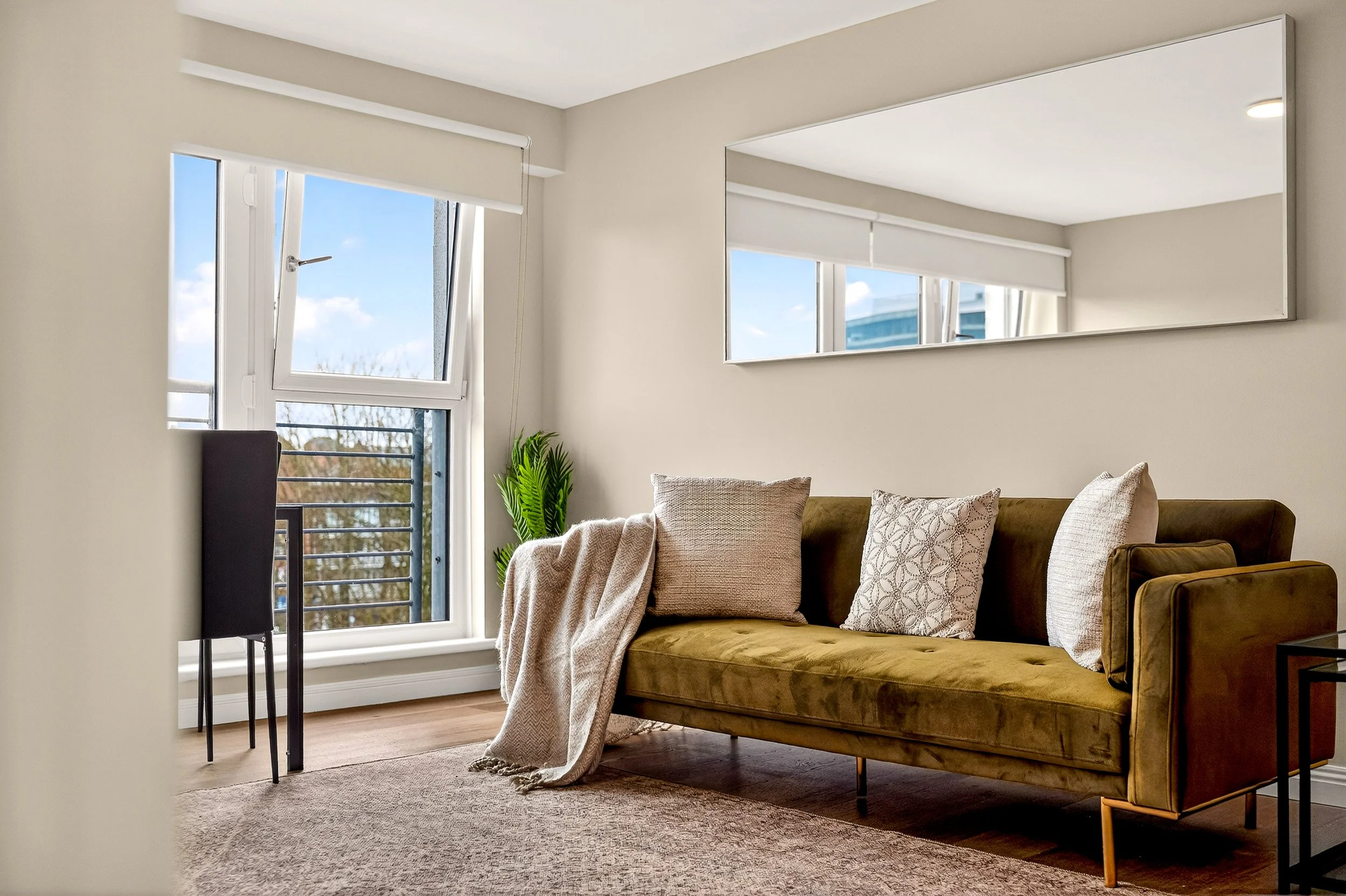 Living room with a green velvet sofa, beige pillows, a beige throw blanket, a black side table, a large window showing a balcony and trees outside, a potted plant, and a rectangular mirror on the wall.