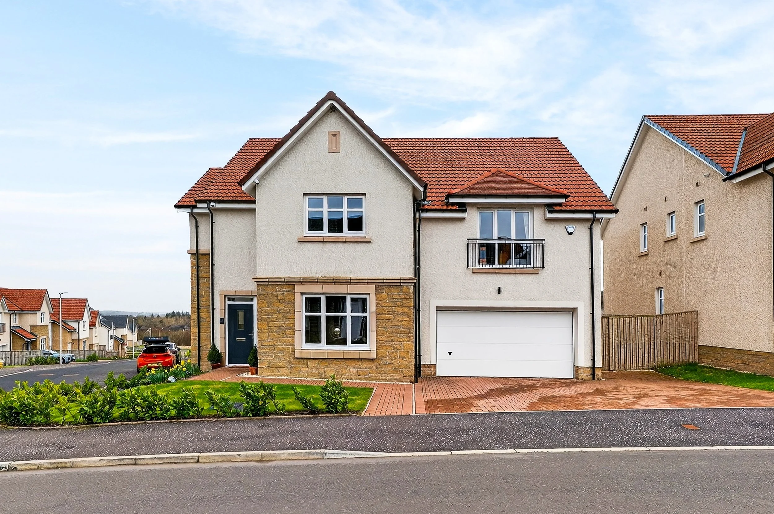 A two-story house with a red tiled roof, beige walls, and a stone facade around the bay window. The house has a white garage door and a small front garden with green plants. There is a driveway and a black car parked on the street in front.