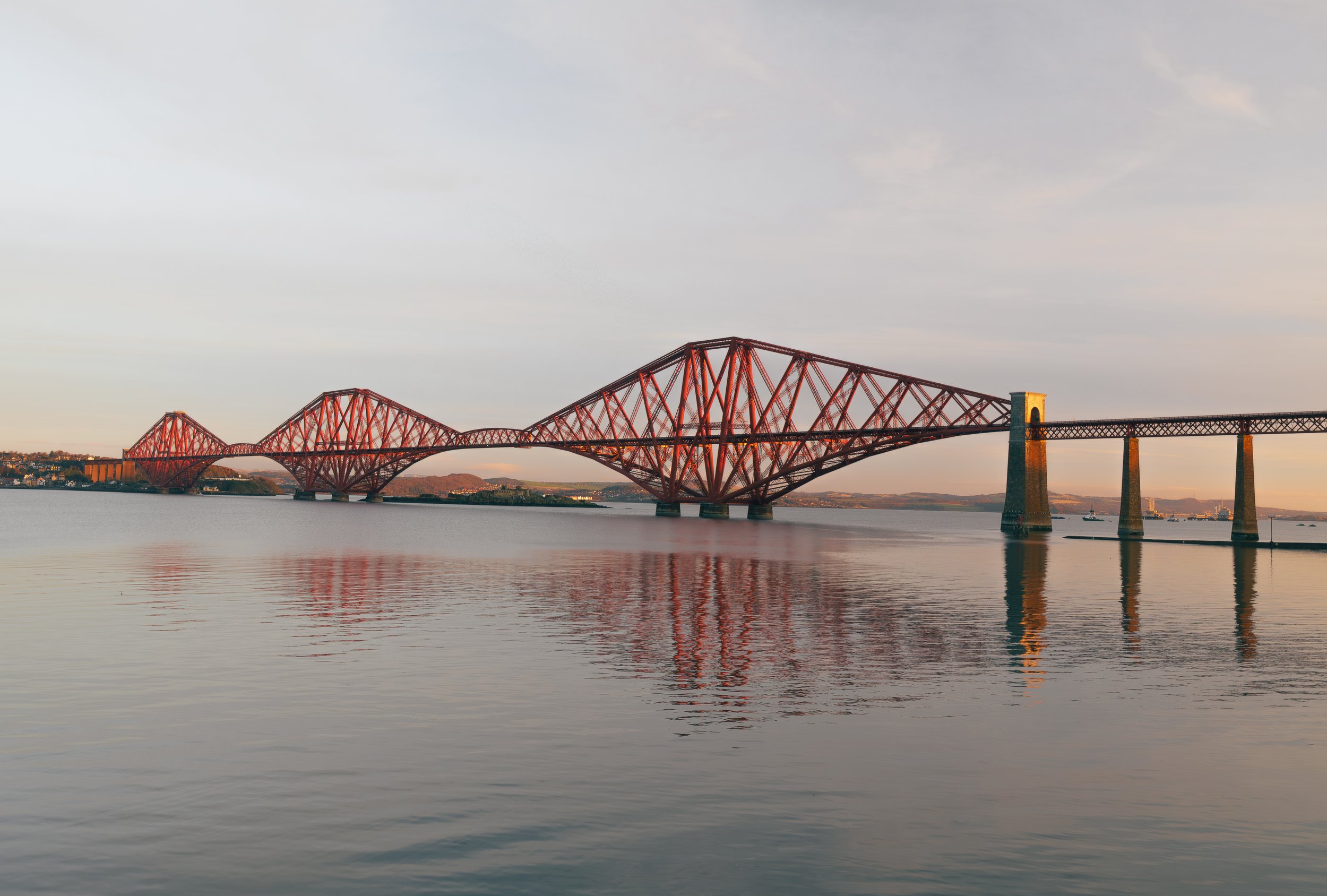 A large red bridge spanning over a body of water during sunset, with reflections visible in the water.