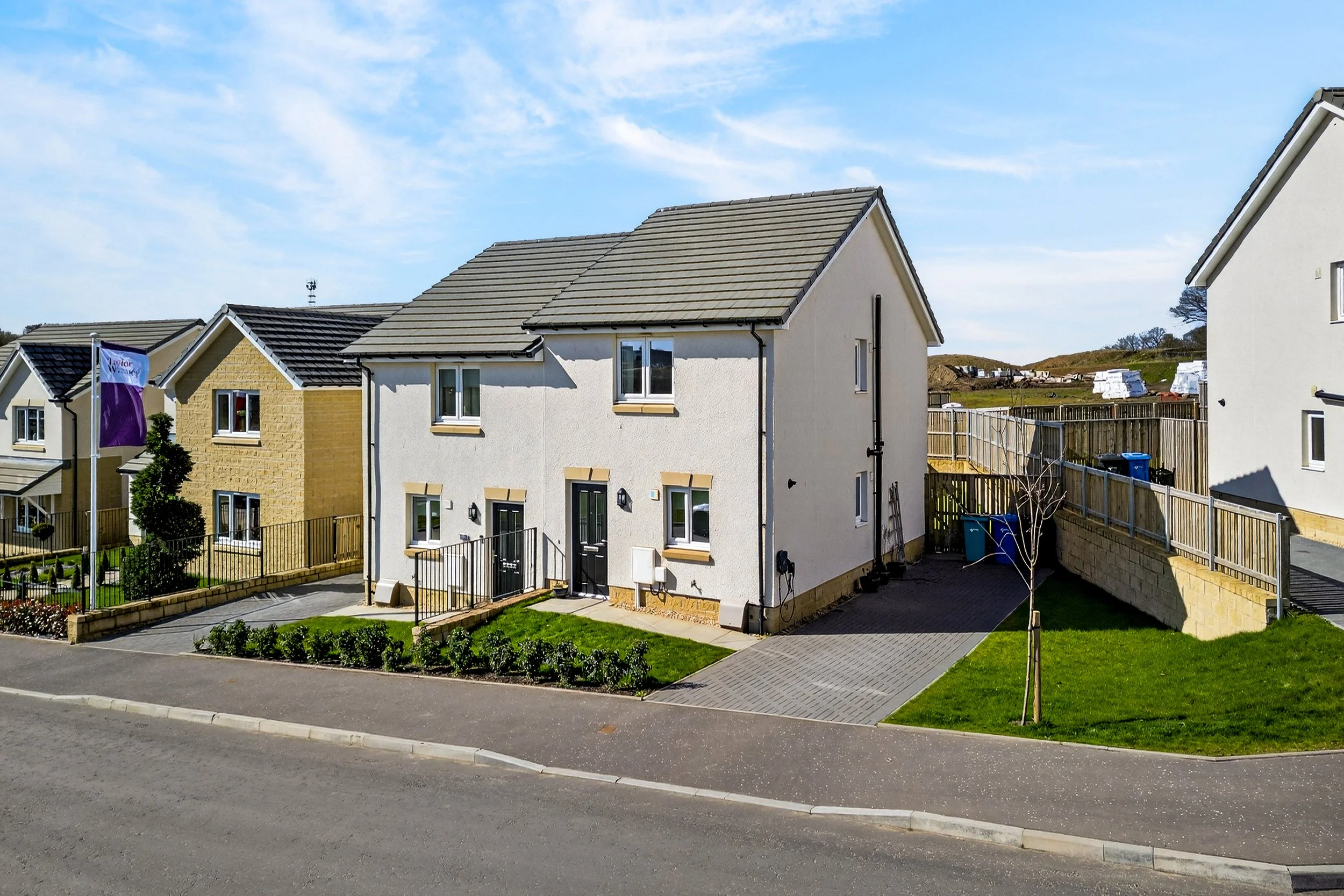 A modern two-story house with a white exterior, black front door, and small front yard with a garden and grass. The house has a driveway on the side, a wooden fence, and neighboring houses visible nearby under a blue sky.