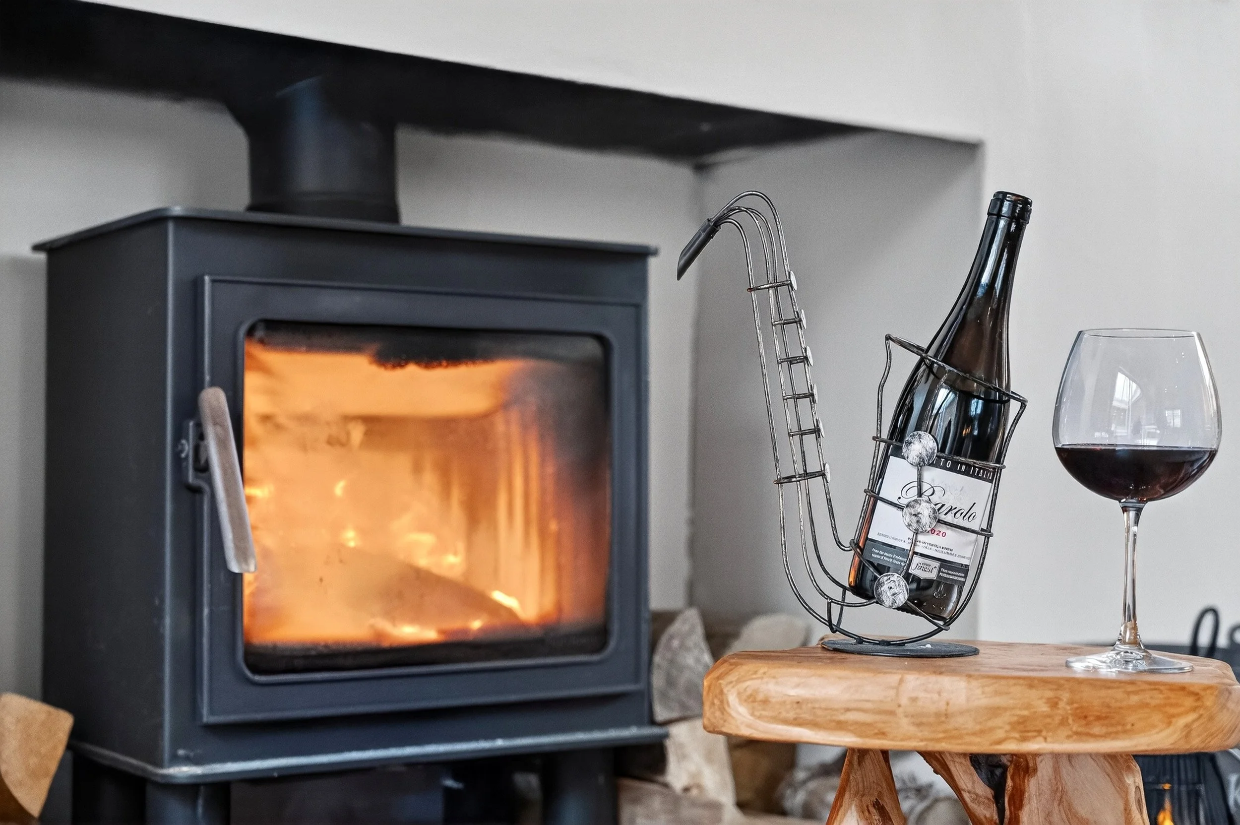 A black wood stove with a glass door showing a burning fire inside, next to a small wooden table holding a wine bottle in a metal wine holder and a glass of red wine.