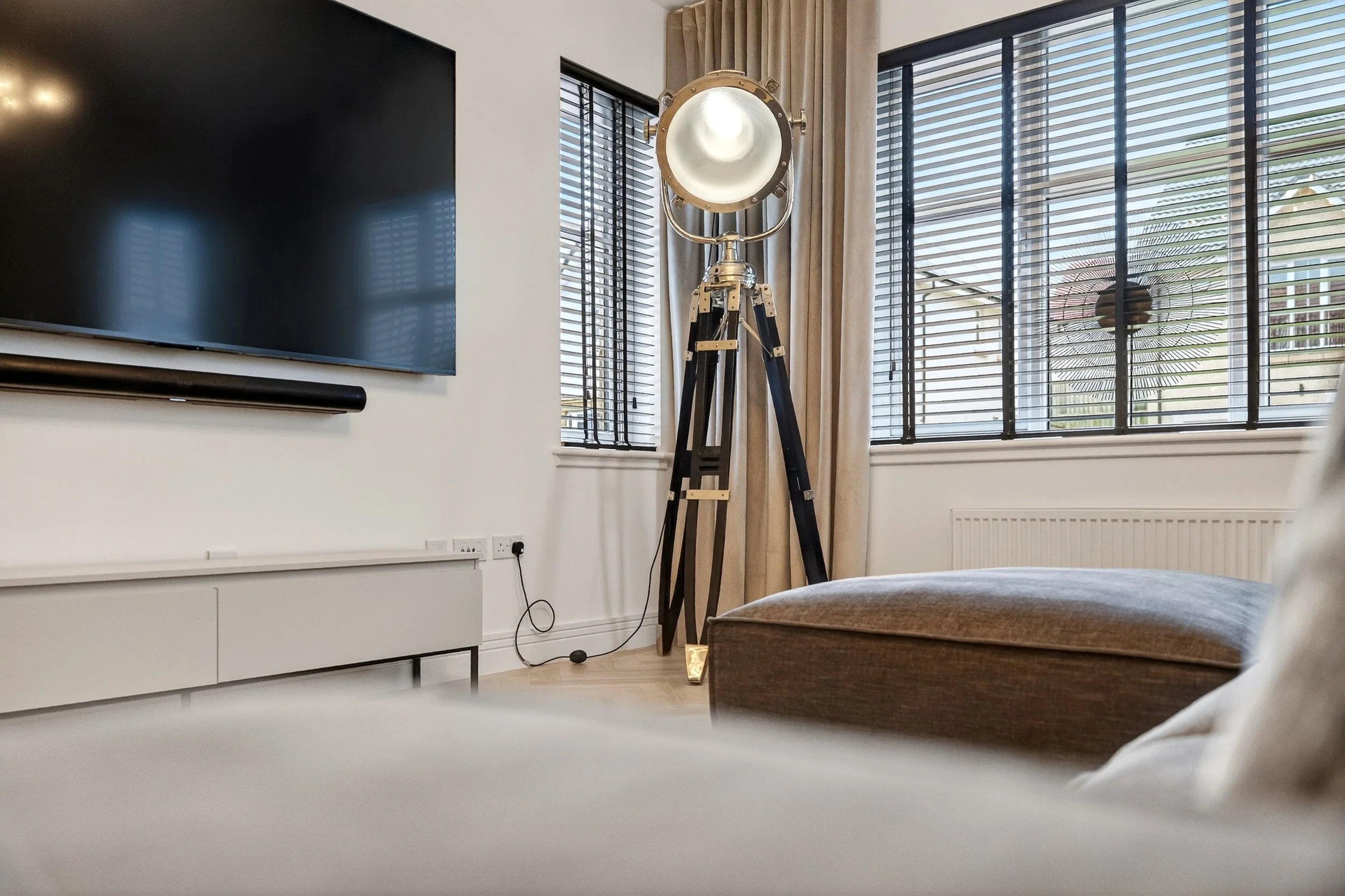 A living room with a large flat-screen TV mounted on the wall, a vintage-style floor lamp resembling a large spotlight, and a window with horizontal blinds and beige curtains.