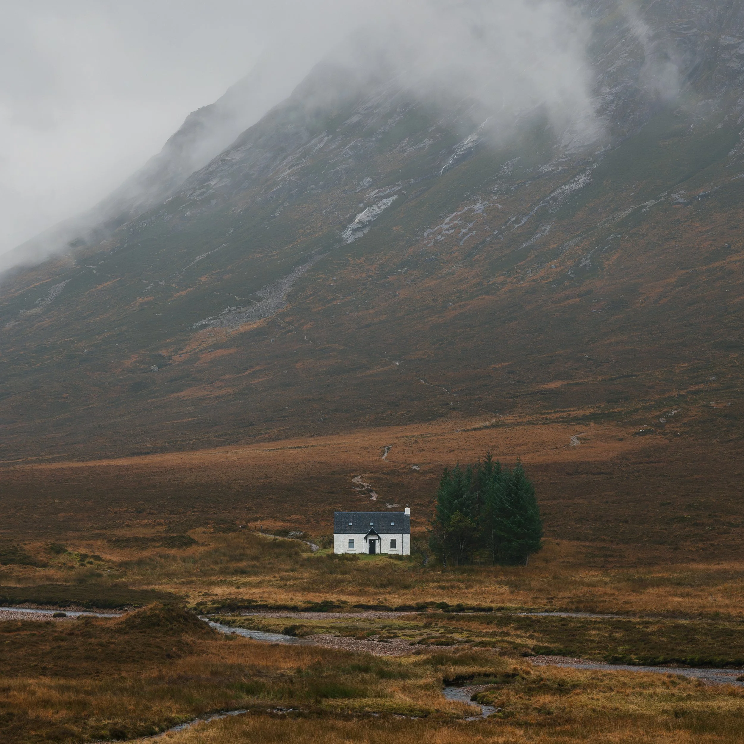 A small white house with a dark roof, situated in a vast, open landscape with mountains in the background. The scene is misty and overcast, with sparse trees nearby and small streams flowing across the grassy terrain.