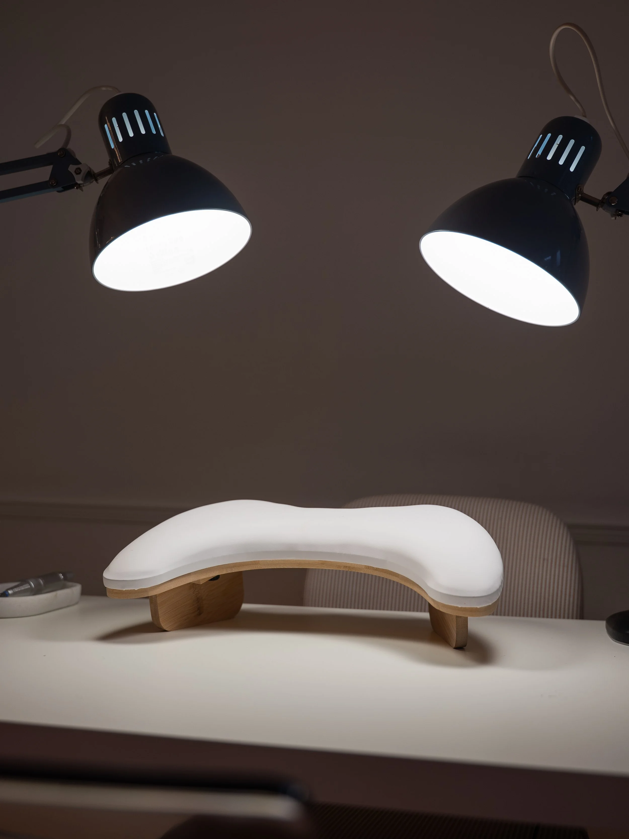 A modern sculptural desk lamp with a white, softly glowing, curved shade and wooden supports, illuminated by two adjustable black and silver adjustable desk lamps above.