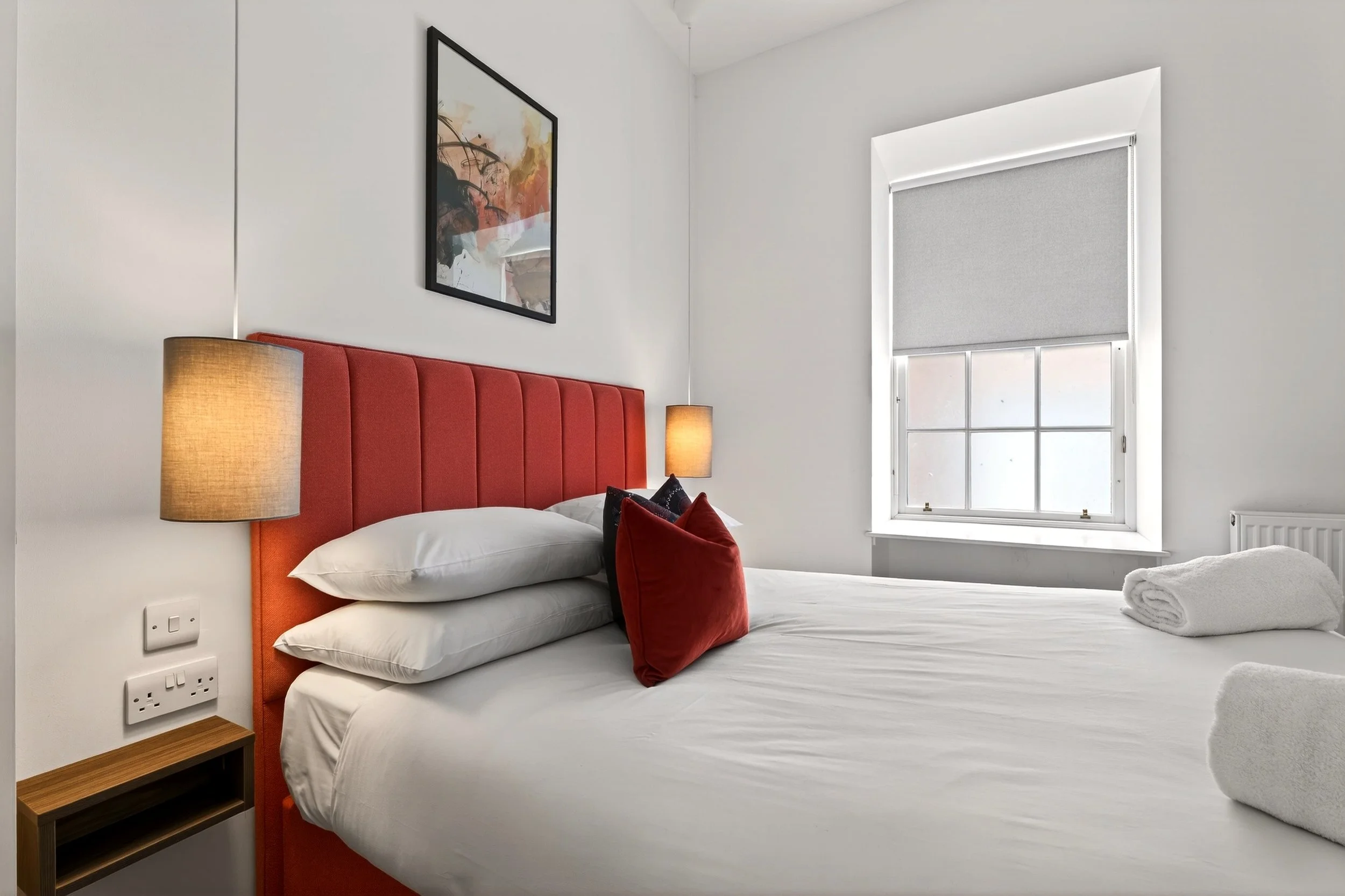 A bedroom with a red upholstered headboard, white bedding, and a decorative red pillow. There are two lamps on either side of the bed, a framed abstract art piece on the wall, and a window with a gray roller shade. Rolled towels are on the bed.