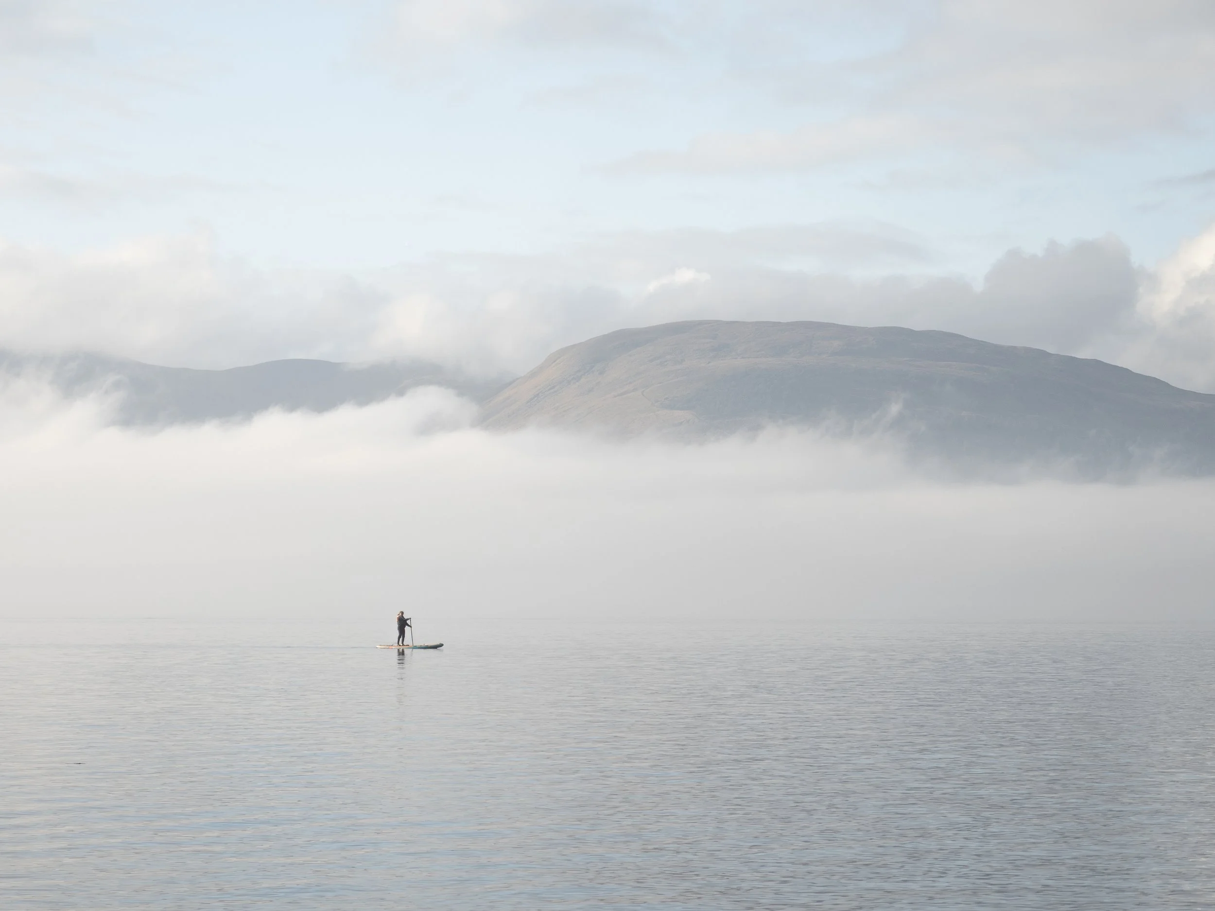 A solitary paddle-boarder on Loch Lomond against a backdrop of fog and hills.