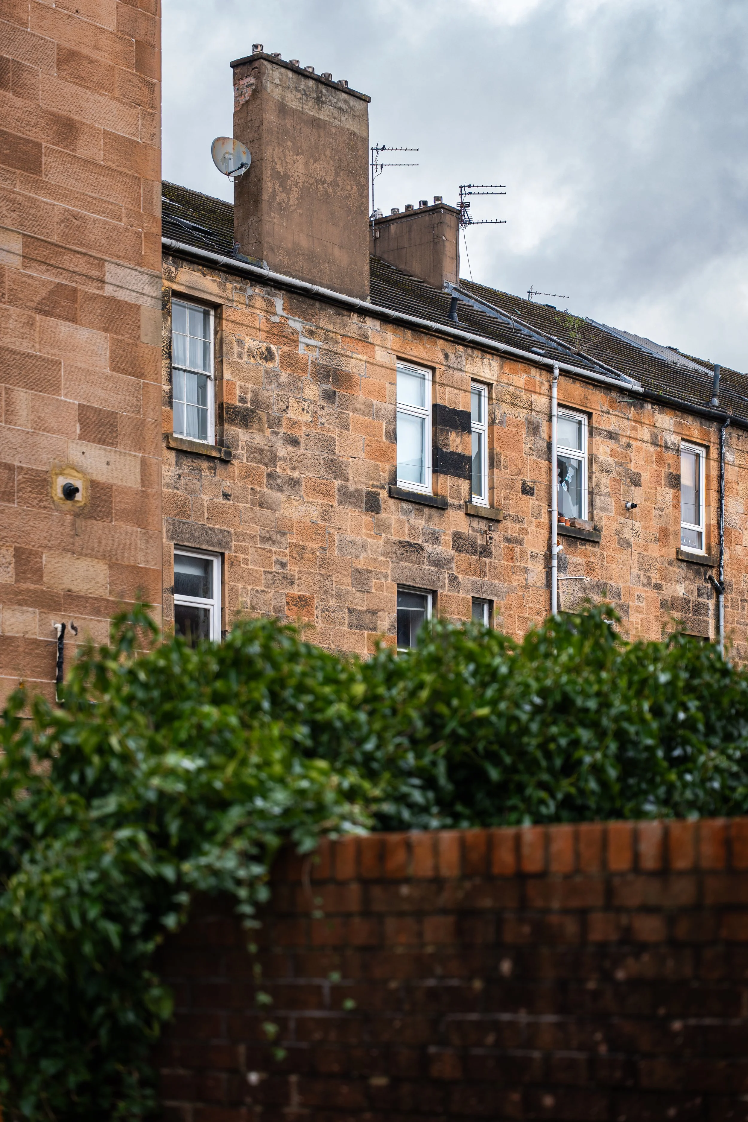 A brick apartment building with multiple windows, chimneys, and TV antennas on the roof, partially obscured by a green hedge and a brick wall in the foreground.