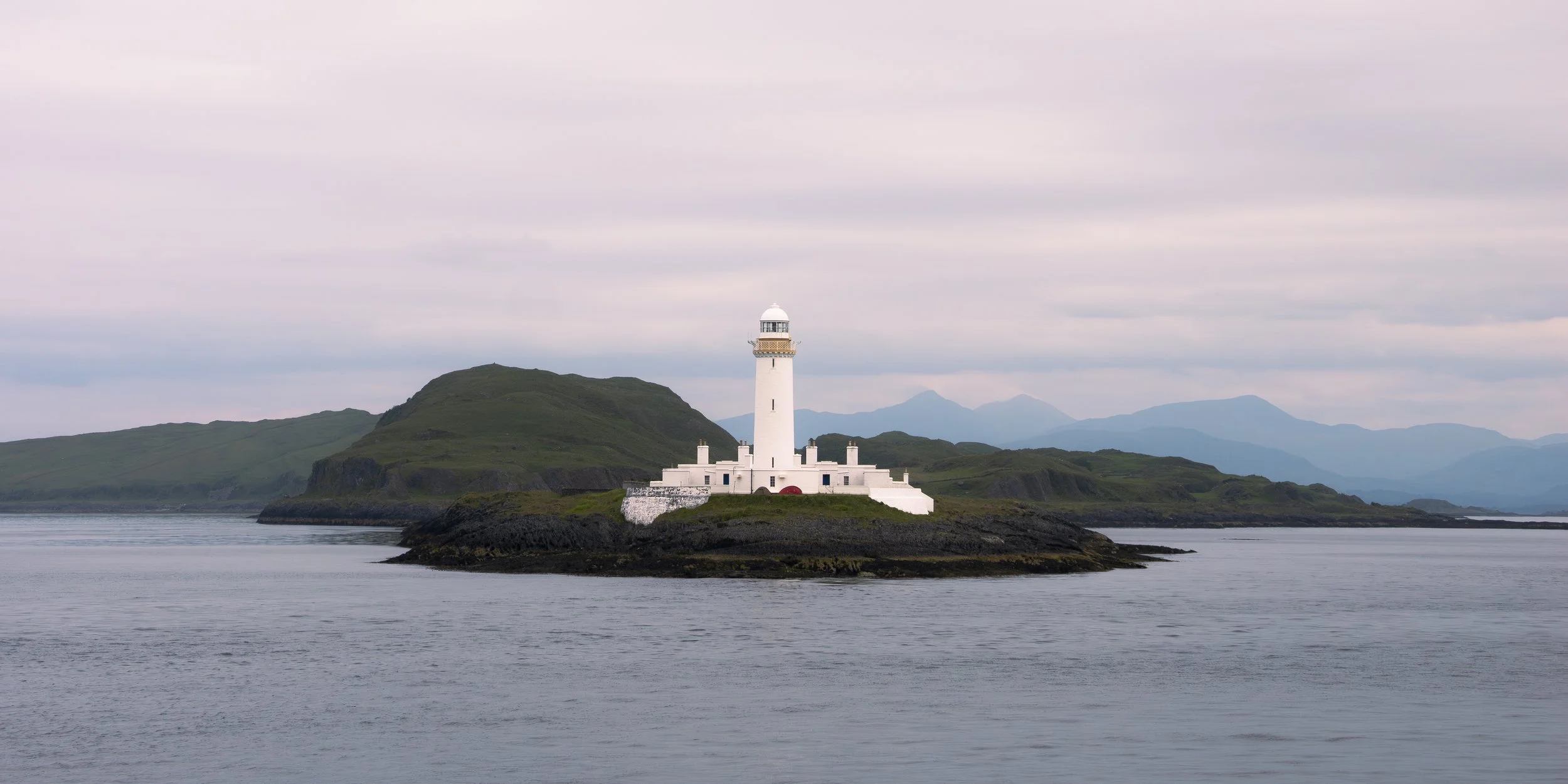 A white lighthouse on a small island surrounded by water, with green hills and mountains in the background on a cloudy day.