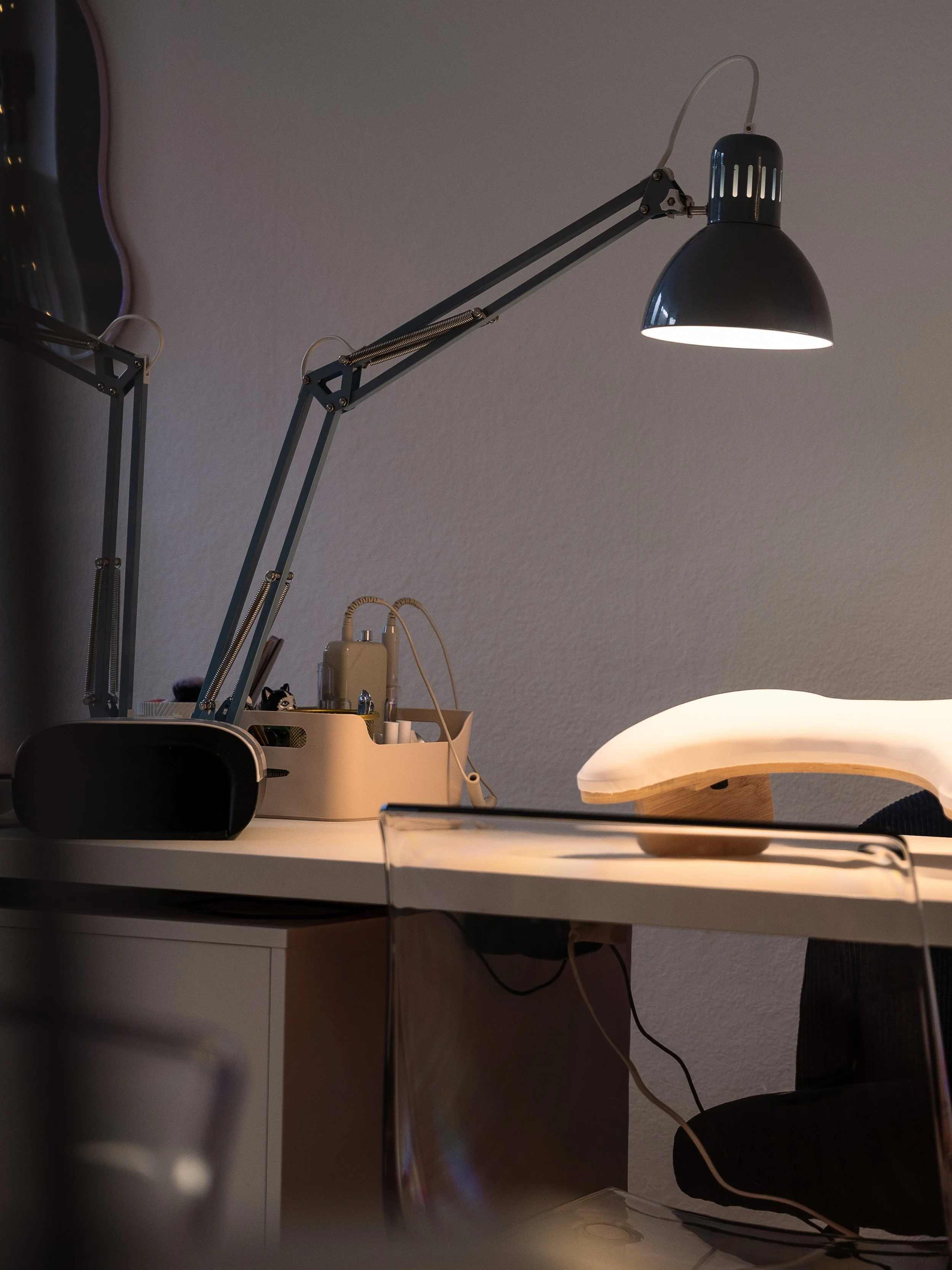 A desk with a black adjustable arm lamp turned on, a lighted curved lamp, and various small items and bottles on the surface.