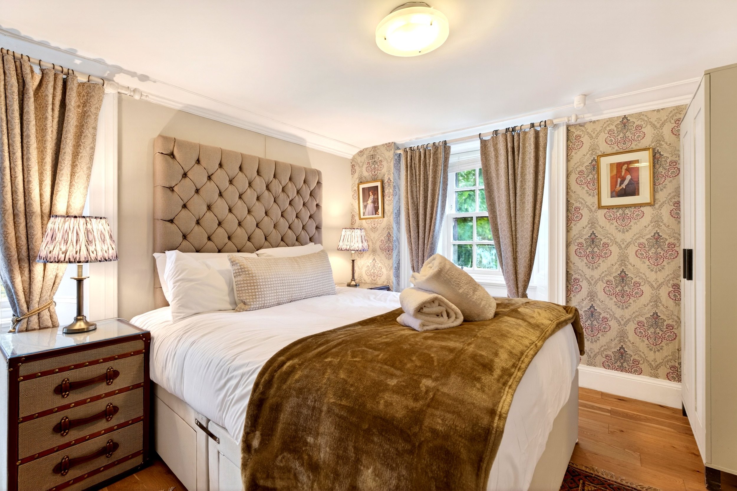 Cozy bedroom with beige tufted headboard, white bed linens, and rolled towels. Gold and patterned curtains, vintage-style bedside lamps, framed artwork on patterned wallpaper, and wooden flooring.