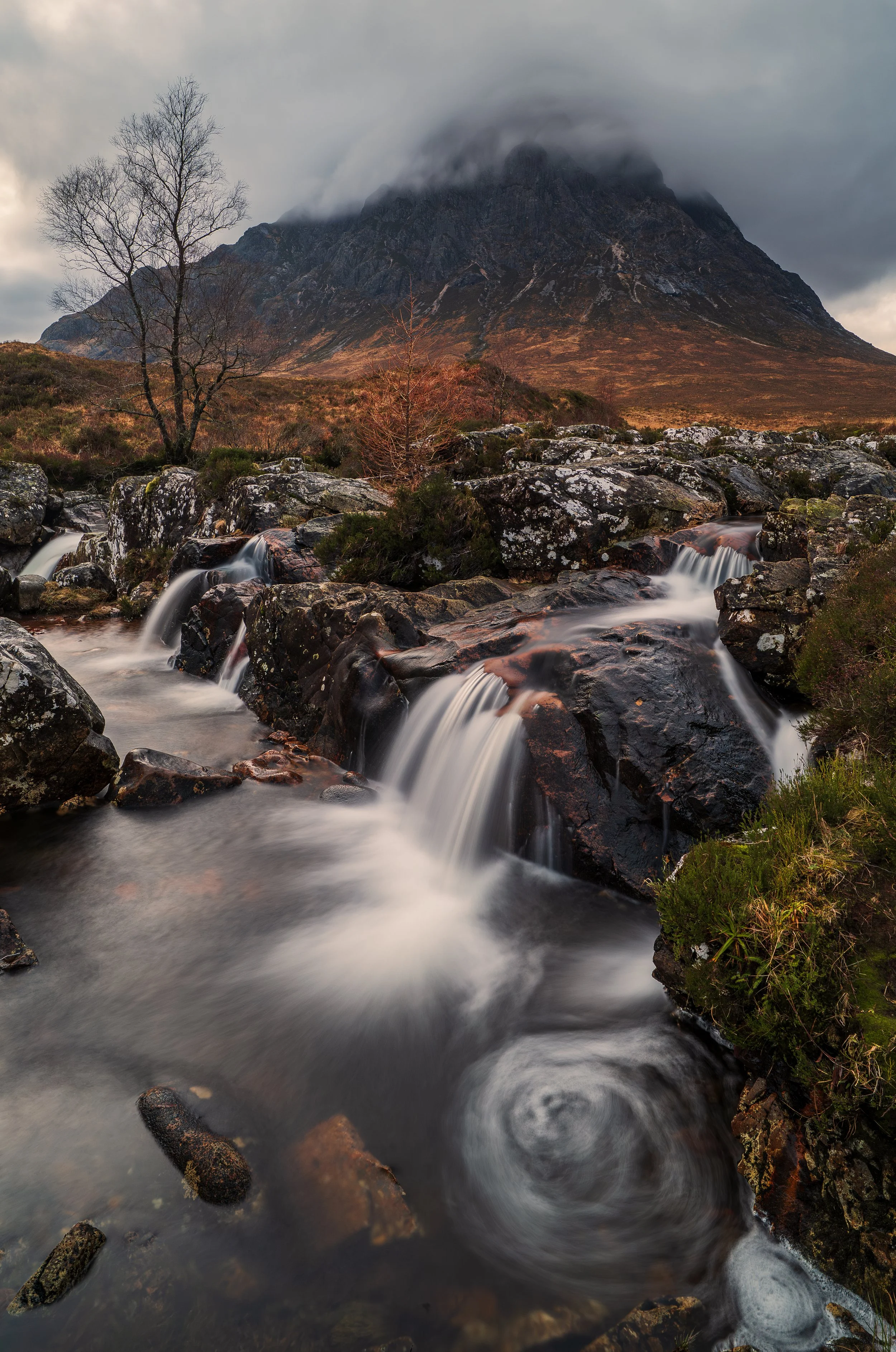 A mountainous landscape featuring a cloudy summit, a flowing stream with small waterfalls, rocks, and sparse trees in a scenic outdoor setting.