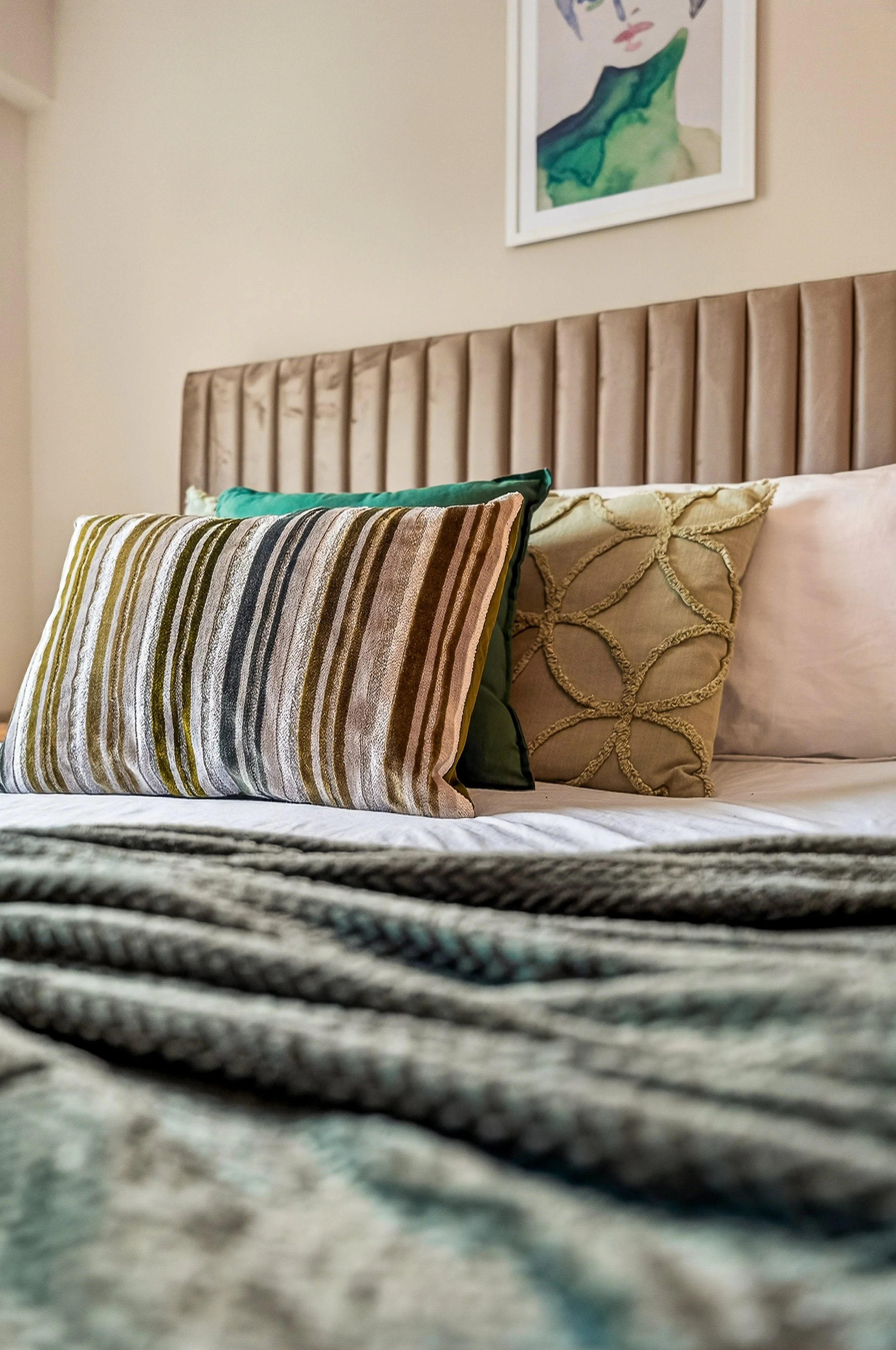 Close-up view of a bed with decorative pillows and a tan tufted headboard, with abstract art on the wall above.