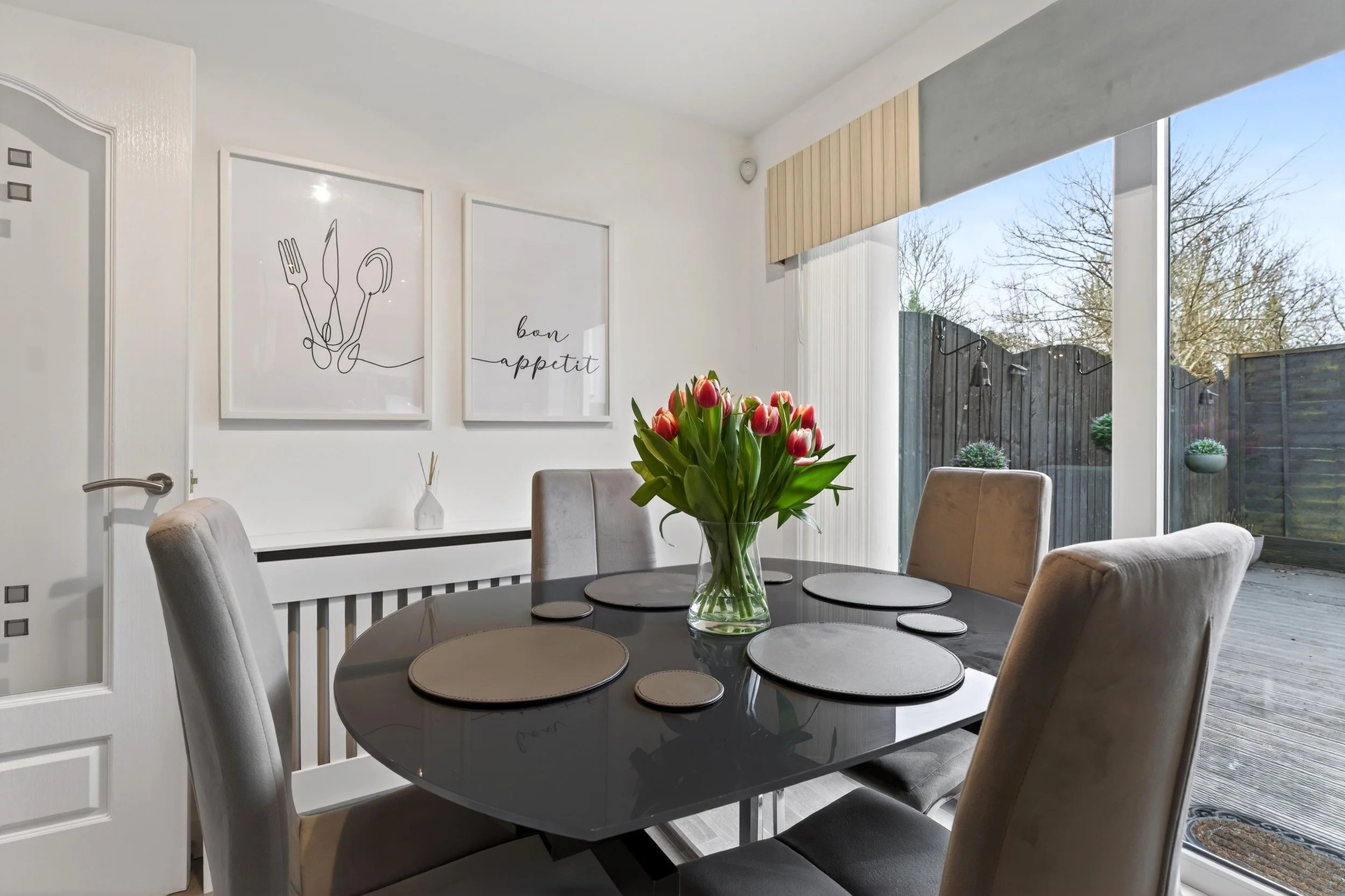 Dining room with a black dining table, beige upholstered chairs, a glass vase with pink tulips, and wall art with cutlery and 'bon appétit' in black lettering.