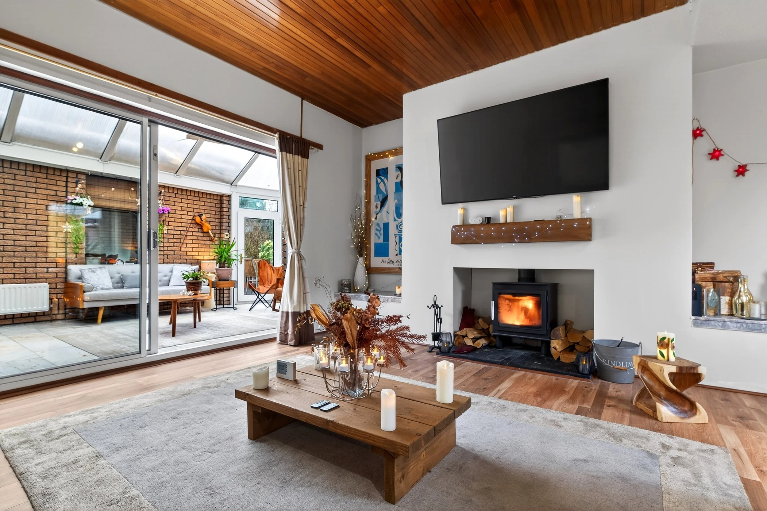 Living room with a wood-burning stove, fireplace logs, a large flat-screen TV mounted on white wall, candles, a wooden mantel, a coffee table with candles and a flower arrangement, and sliding glass doors leading to an enclosed patio with outdoor sea