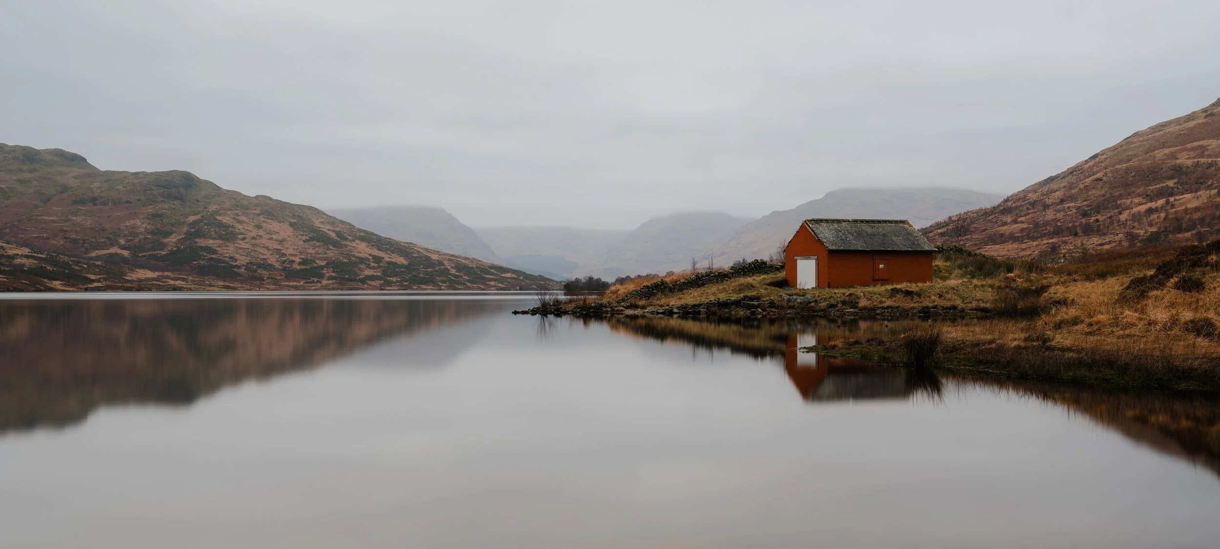A grey, still day on Loch Arklet, with reflections of a small red hut and the hills in the water.