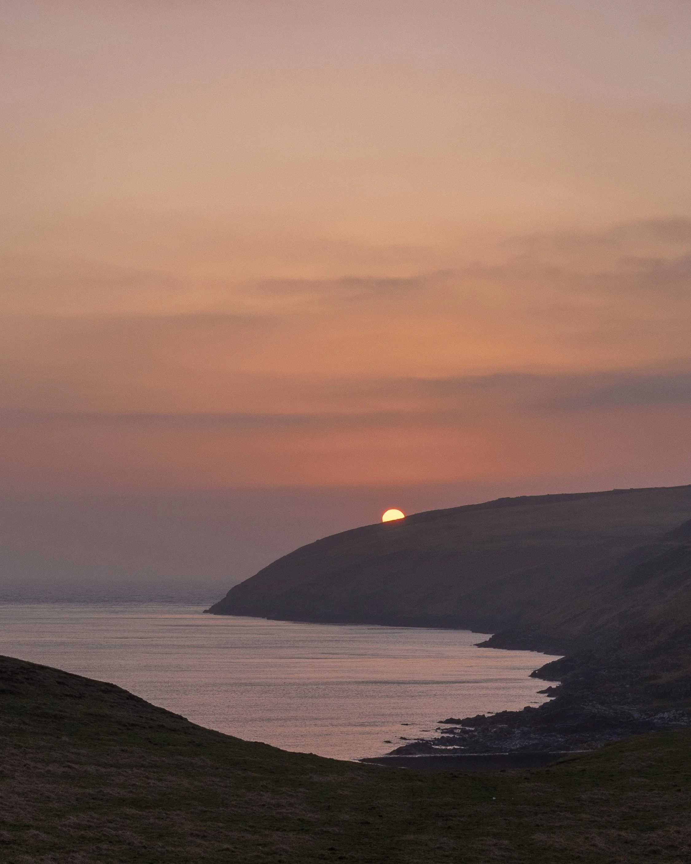Sunset over a coastal landscape with rolling green hills and calm water.