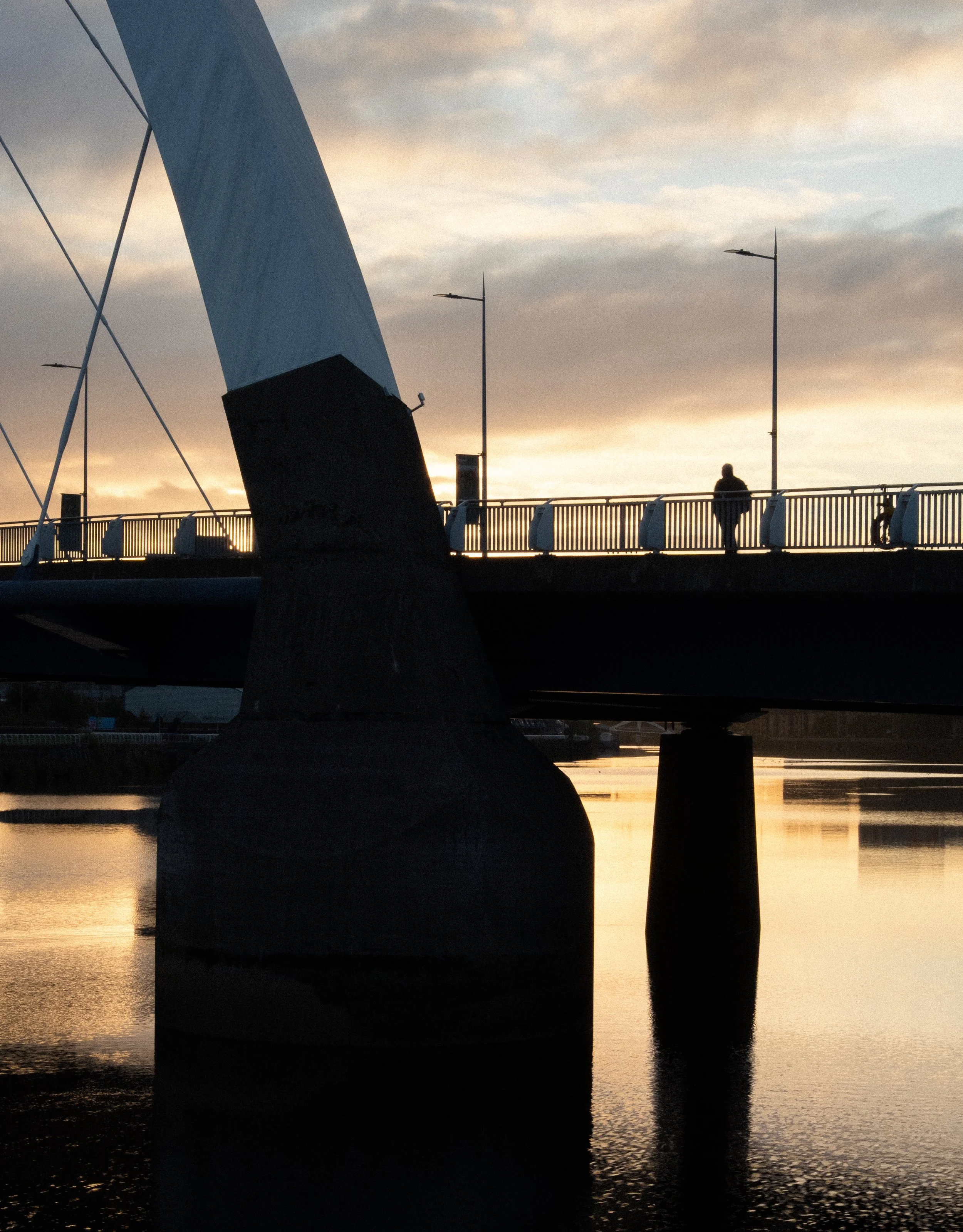 A person walking on a bridge at sunset, with the bridge's support pillar and water below reflecting the sky.