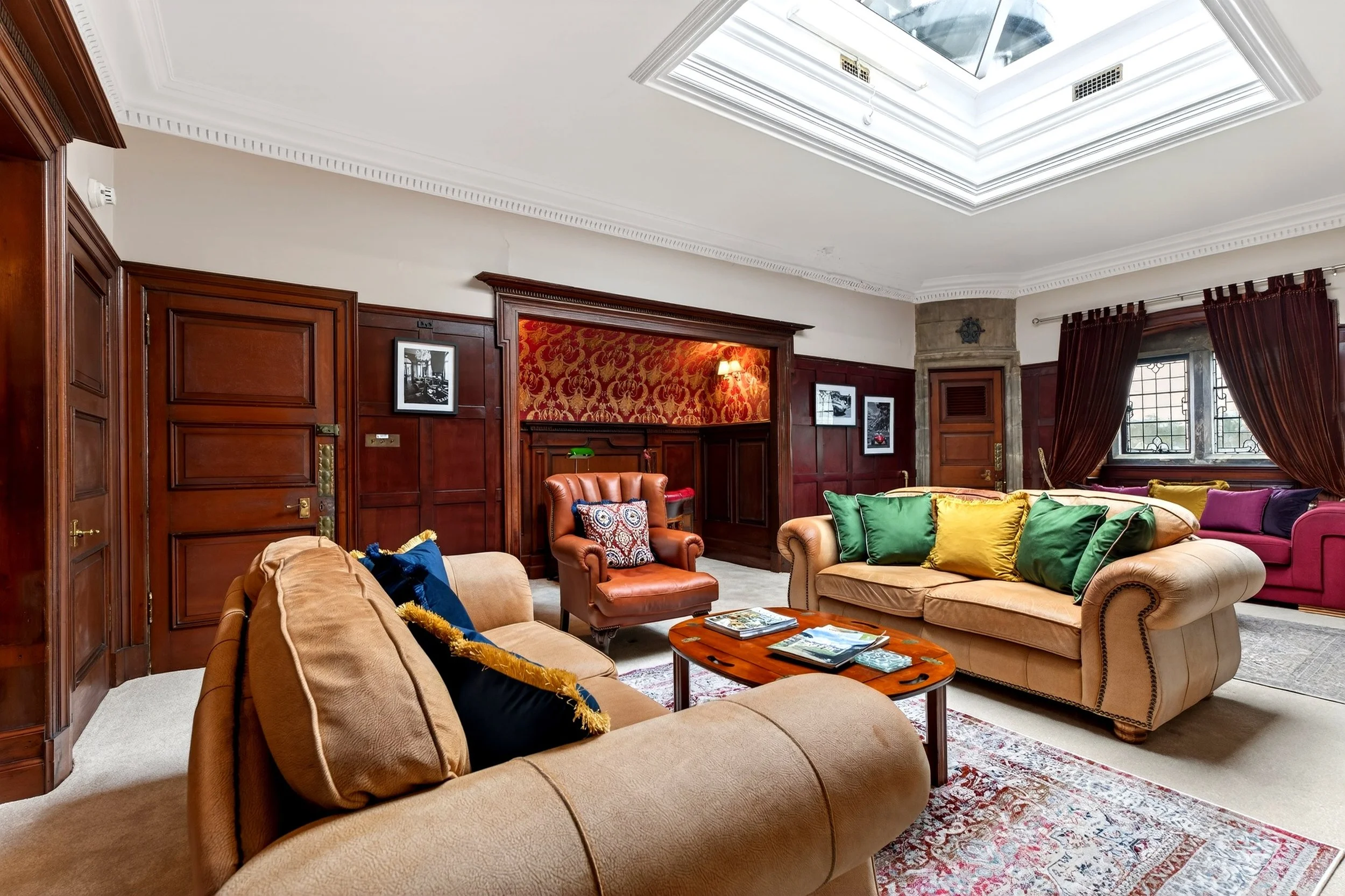 Elegant living room with wooden panel walls, multiple sofas with colorful cushions, a wooden coffee table with magazines, a window with stained glass and curtains, and a skylight in the ceiling.
