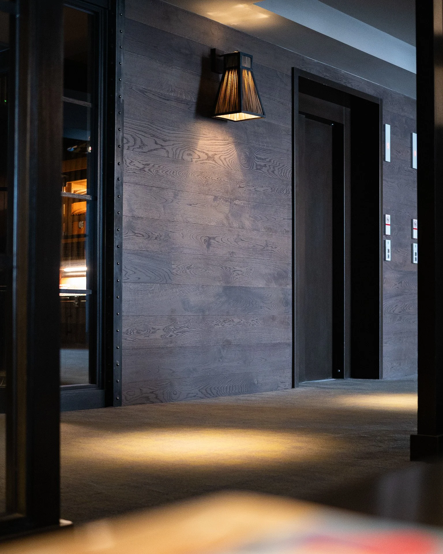 Interior of a modern building with two elevator doors, a wooden wall, and a wall-mounted lamp illuminating the space.
