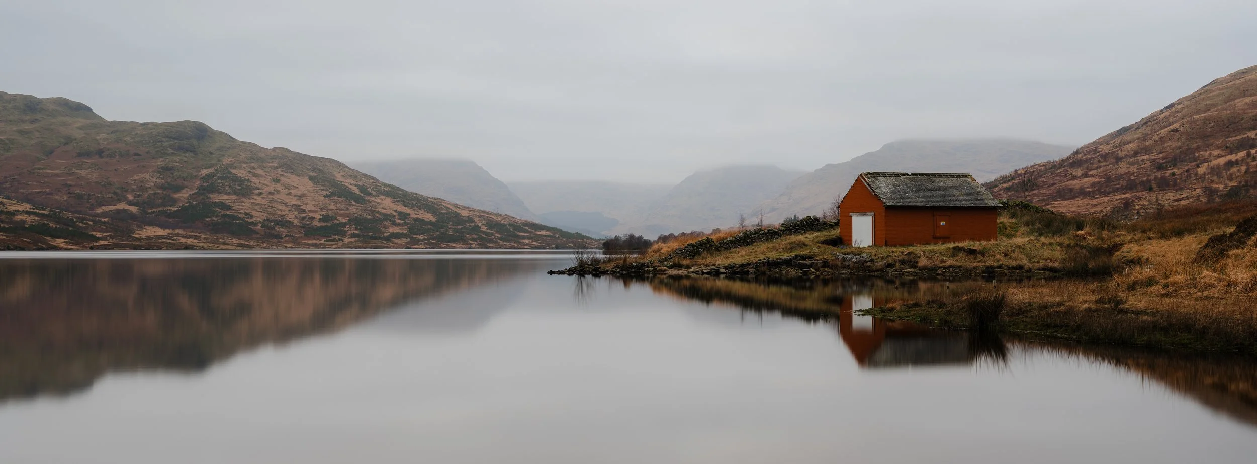 A still, grey day on Loch Arklet, with reflections of a small red hut and the hills in the water.