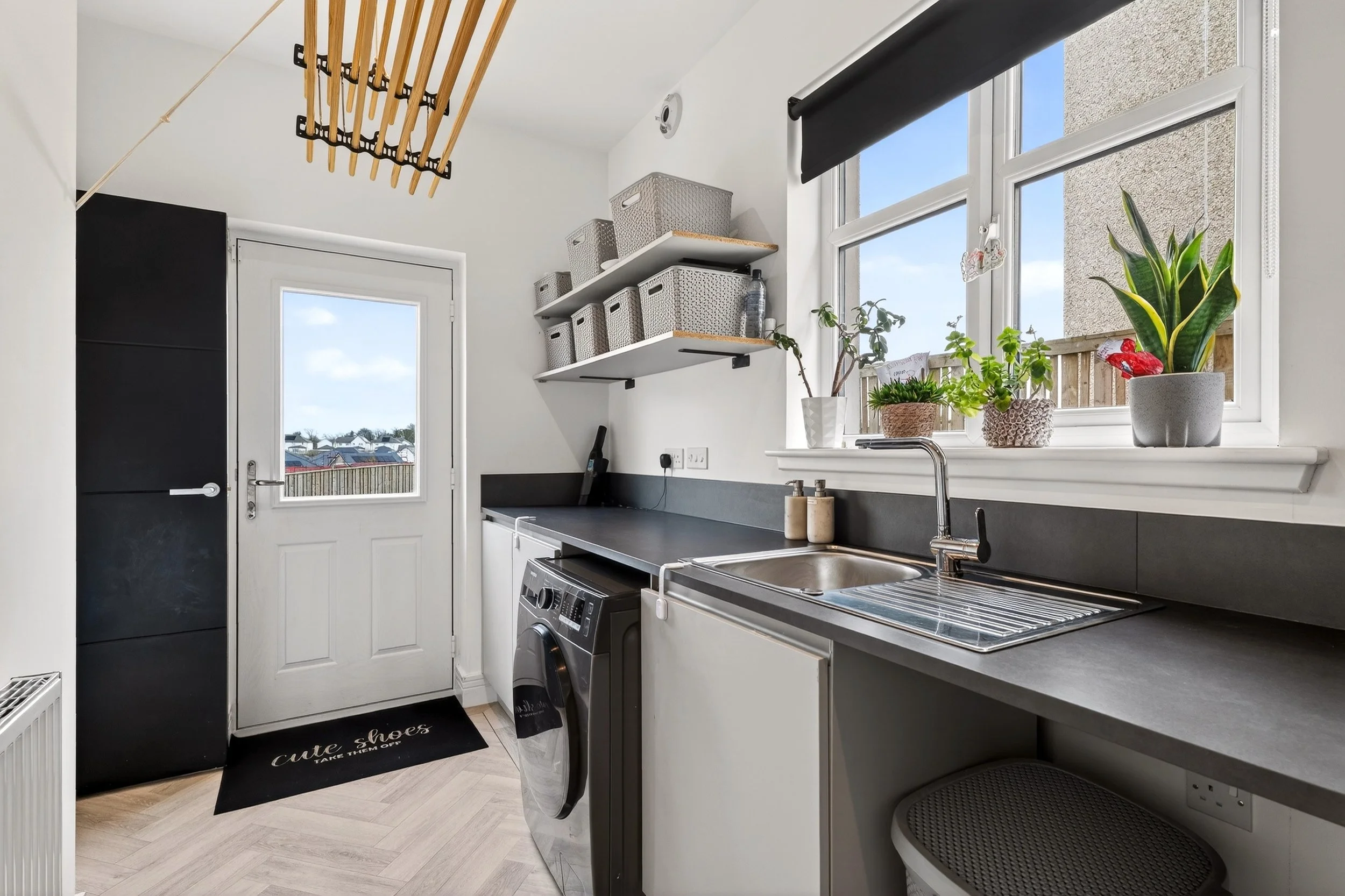 Modern laundry room with black countertop, white cabinets, a washing machine, potted plants on the window sill, and a door leading outside, with shelves holding storage baskets.