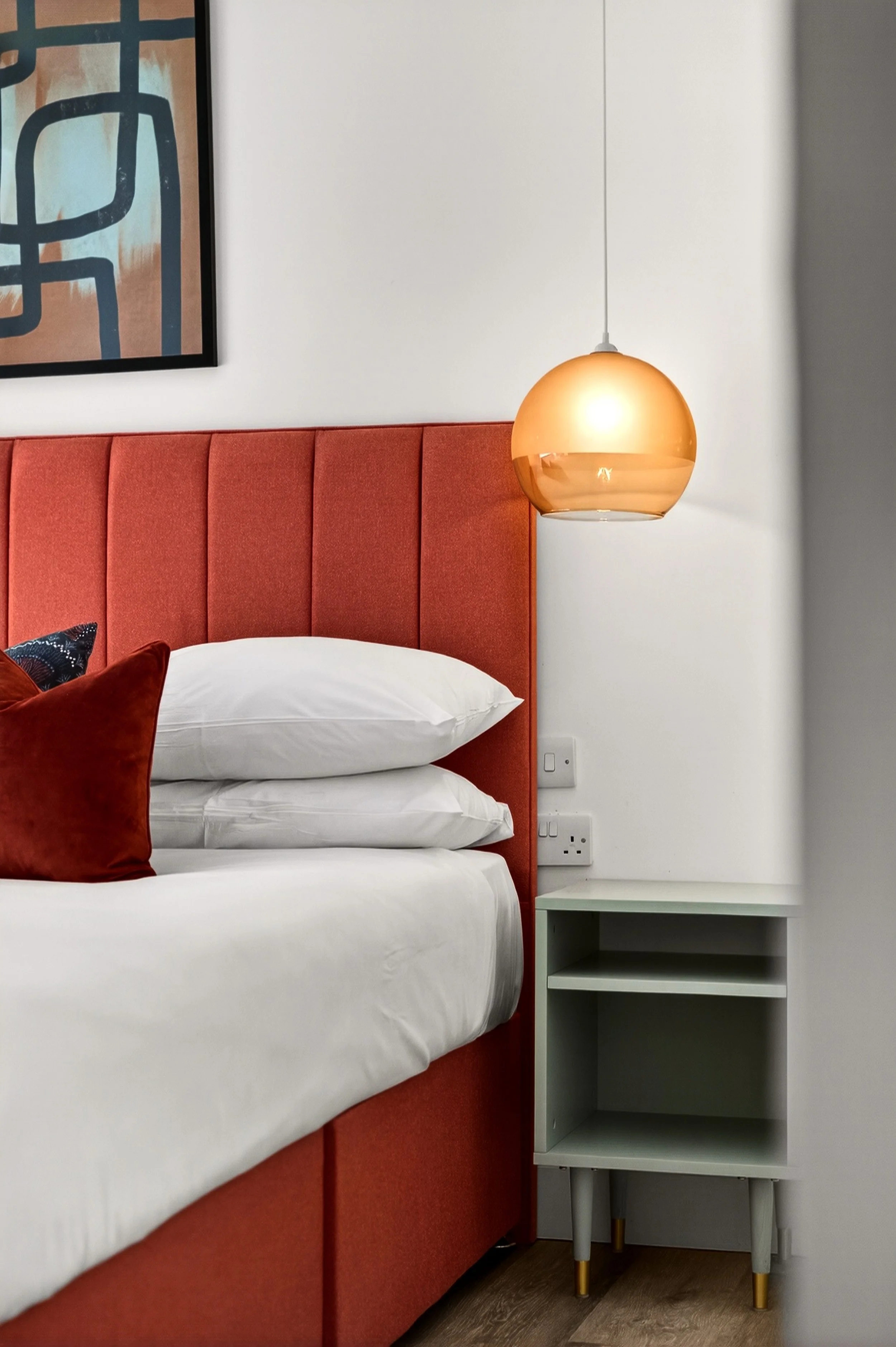 Close-up of a bedroom corner with a red upholstered headboard, a white bed with pillows and a velvet red pillow, a modern hanging amber pendant light, and a small light green nightstand with gold-tipped legs.