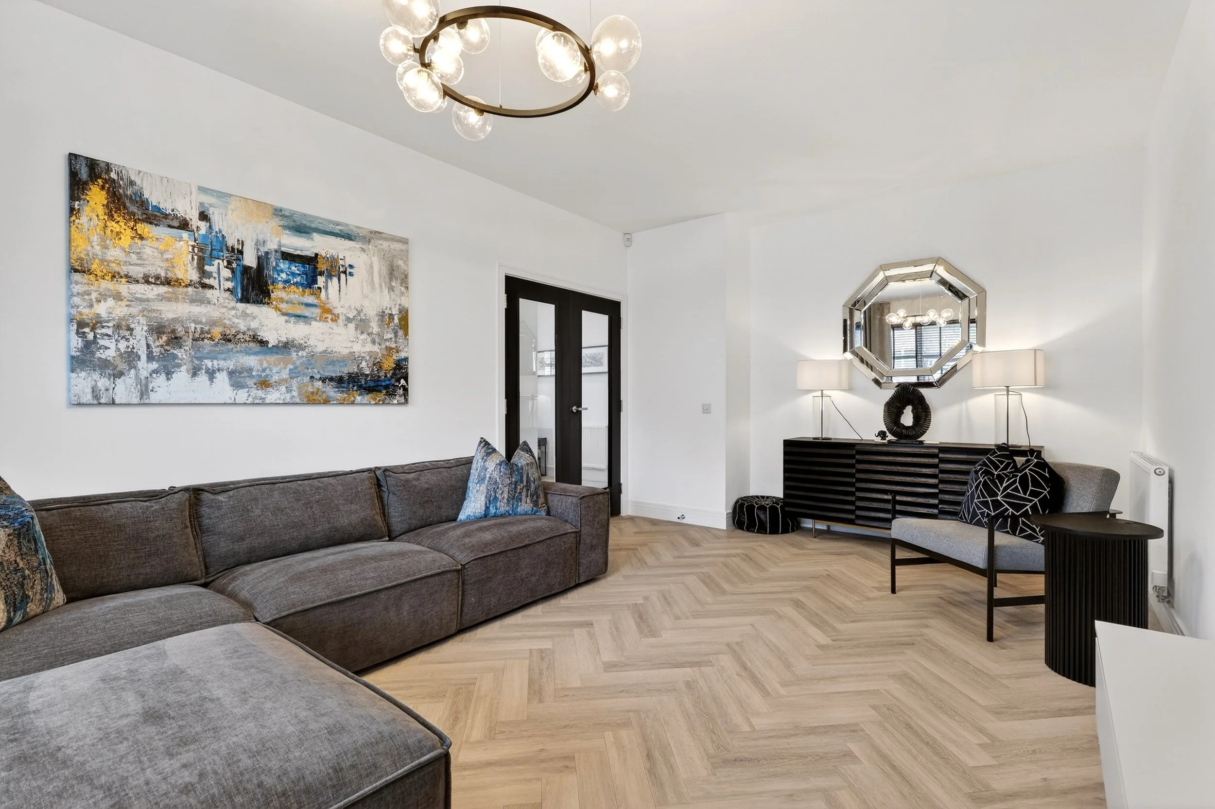 Modern living room with gray sectional sofa, abstract artwork on white wall, decorative pillows, black sideboard with lamps and mirror, gray armchair, black side table, wooden chevron floor, and black door.