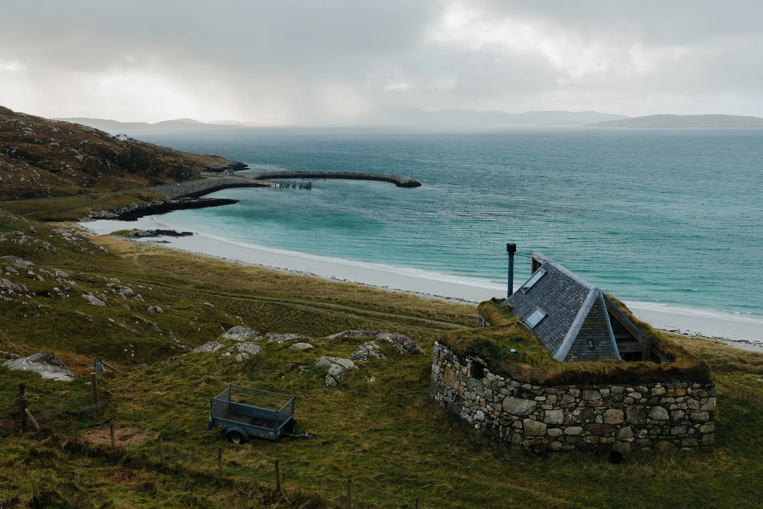 A small stone house with an angled roof and chimney on a grassy hillside overlooking a beach and ocean, with a walking trail, fence, and a trailer in the foreground.